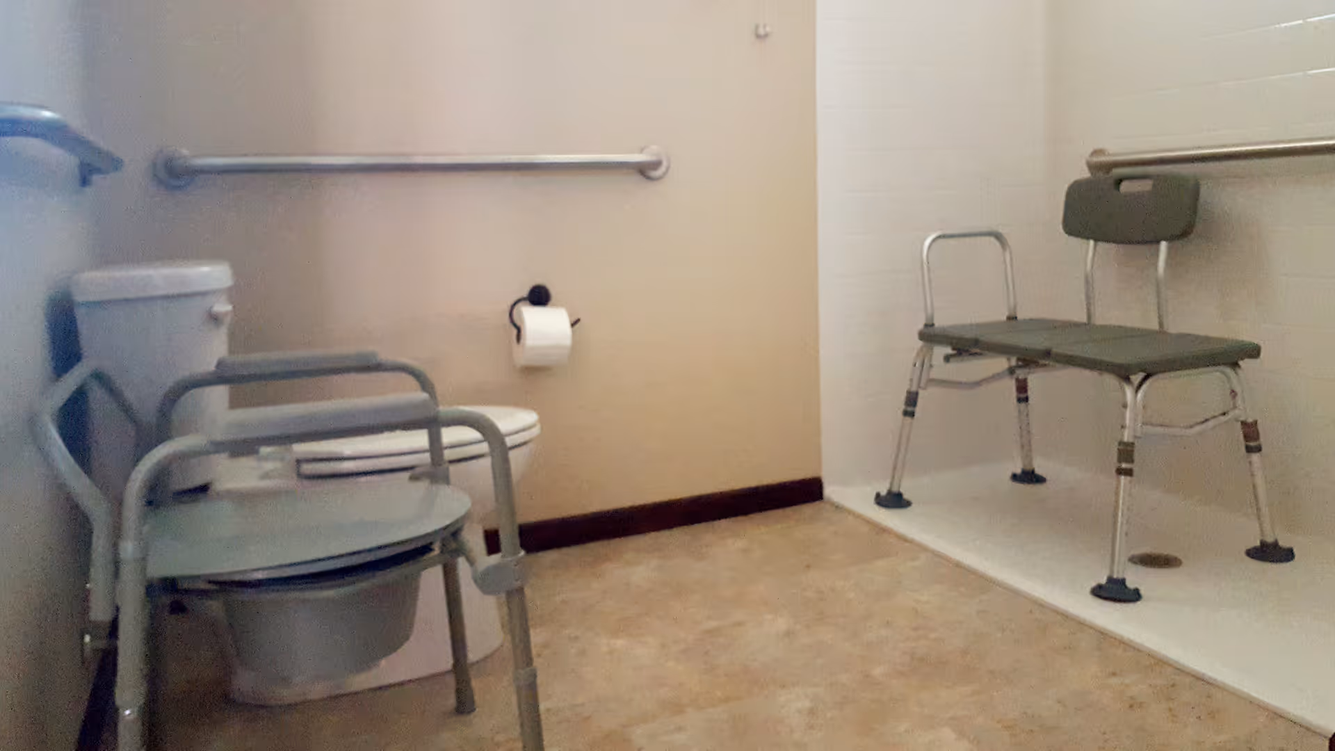 Accessible bathroom with a toilet, portable commode chair, shower bench and grab bars.