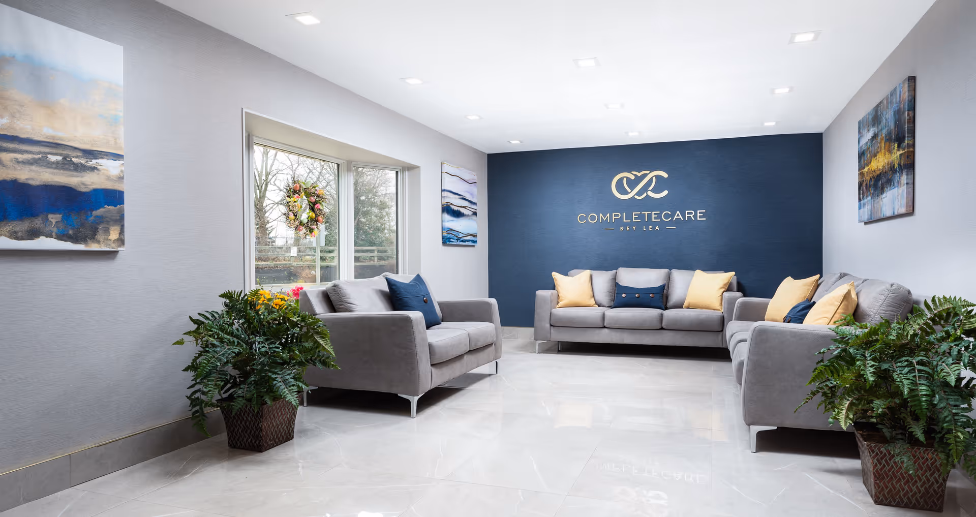 A modern and clean lounge area with three gray sofas arranged around the room, decorated with yellow and blue cushions. The back wall is painted dark blue with the Complete Care at Bey Lea logo and name displayed in gold. There are large windows with a wreath, abstract paintings on the walls, and potted plants on the floor.