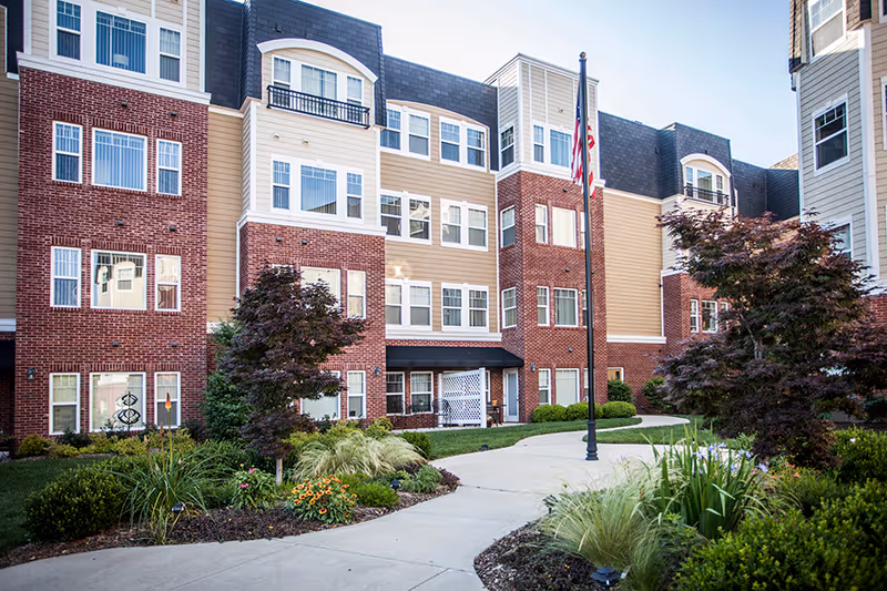 Exterior view of a multi-story senior living facility building with a combination of red brick and beige siding. The building has numerous windows and a landscaped garden with shrubs, flowers, and small trees along a curved concrete pathway. An American flag is displayed on a flagpole near the center of the image.