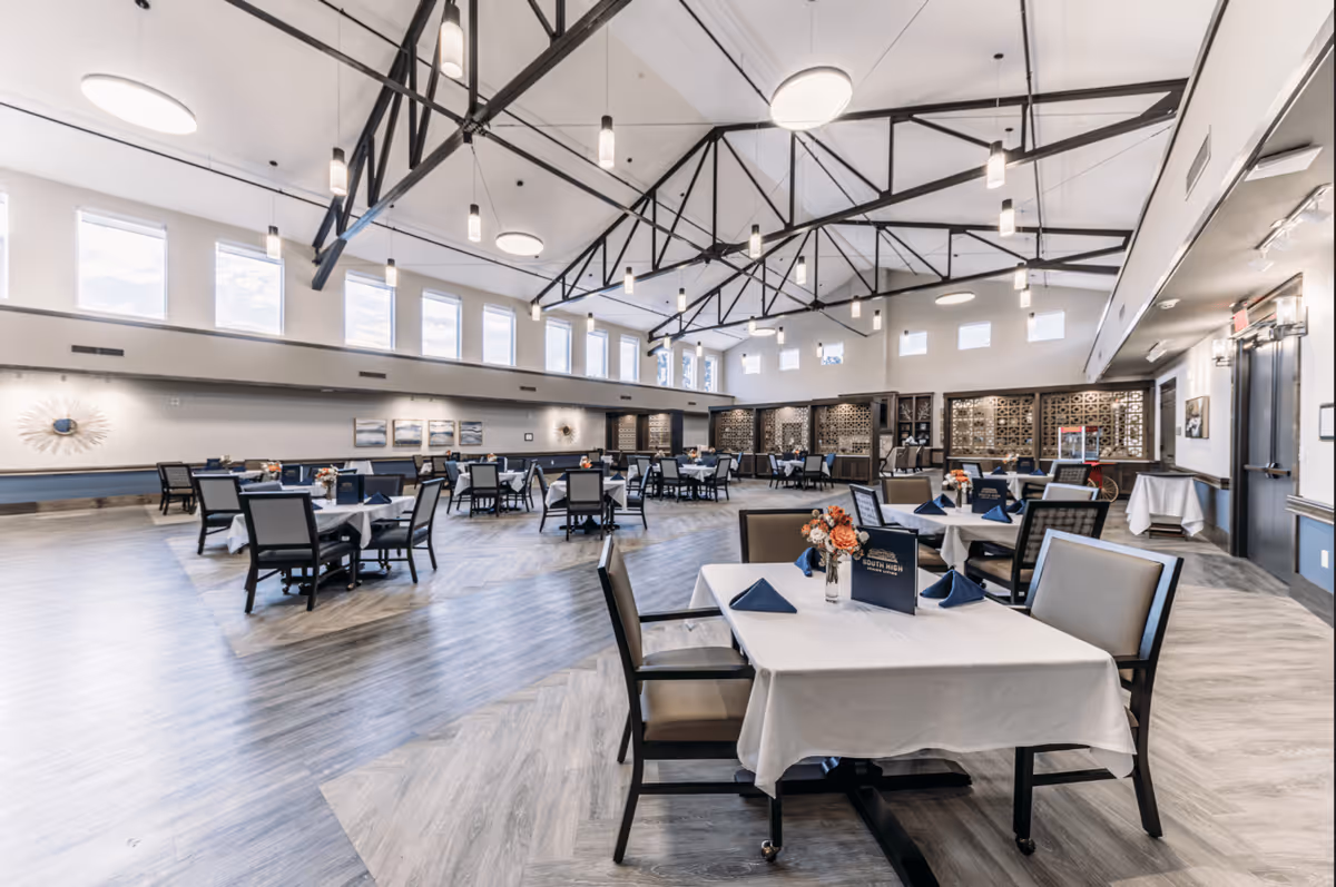 A spacious and well-lit dining room with multiple tables covered in white tablecloths, each set with blue napkins and floral centerpieces. The room features high ceilings with exposed black metal beams and numerous hanging pendant lights. Large windows near the ceiling allow natural light to fill the space. The floor has a wood-like pattern, and the walls are decorated with artwork and decorative panels.
