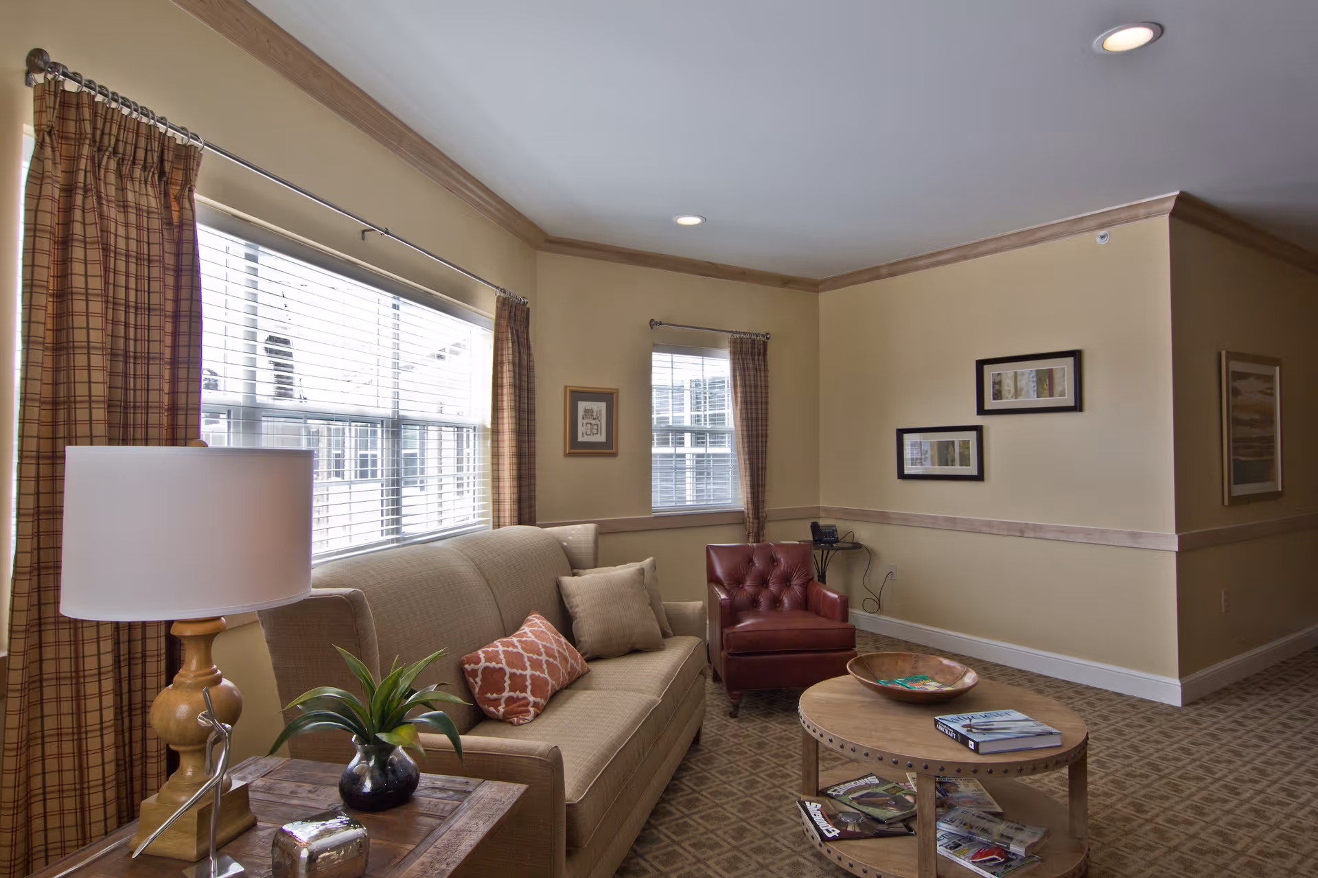 A cozy living room with a beige sofa adorned with two pillows, a red leather armchair, a wooden coffee table with magazines and a decorative bowl, a side table with a lamp and a small plant, beige walls with framed artwork, and windows with plaid curtains letting in natural light.