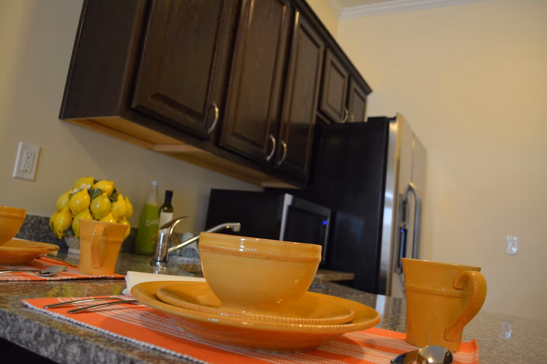 Kitchen countertop set with orange dishware and placemats, sink and faucet, dark wooden cabinets and a stainless refrigerator in the background.