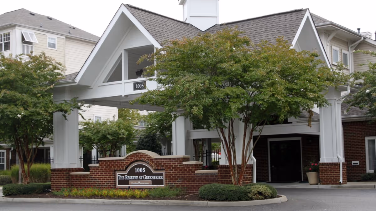 Entrance to The Reserve at Greenbrier facility featuring a covered driveway with white pillars and a peaked roof. There are trees and shrubs around the entrance, and a brick sign displaying the address 1005 and the facility name.
