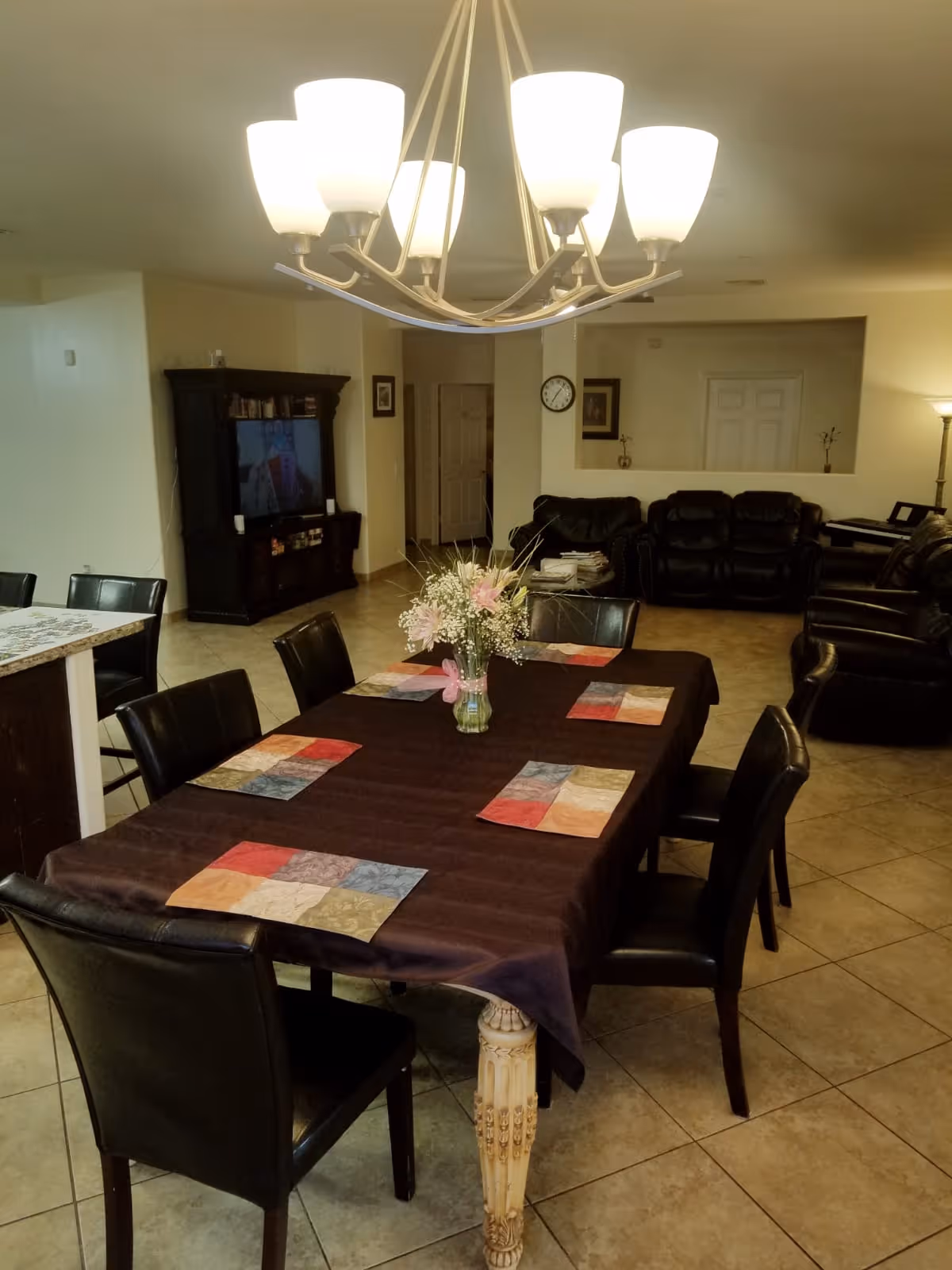 Interior view of a senior living facility showing a dining area with a table covered by a dark tablecloth and colorful placemats, surrounded by black chairs. A vase with flowers is placed in the center of the table. In the background, there is a living room area with black leather sofas, a TV on a wooden stand, a clock on the wall, and a floor lamp.