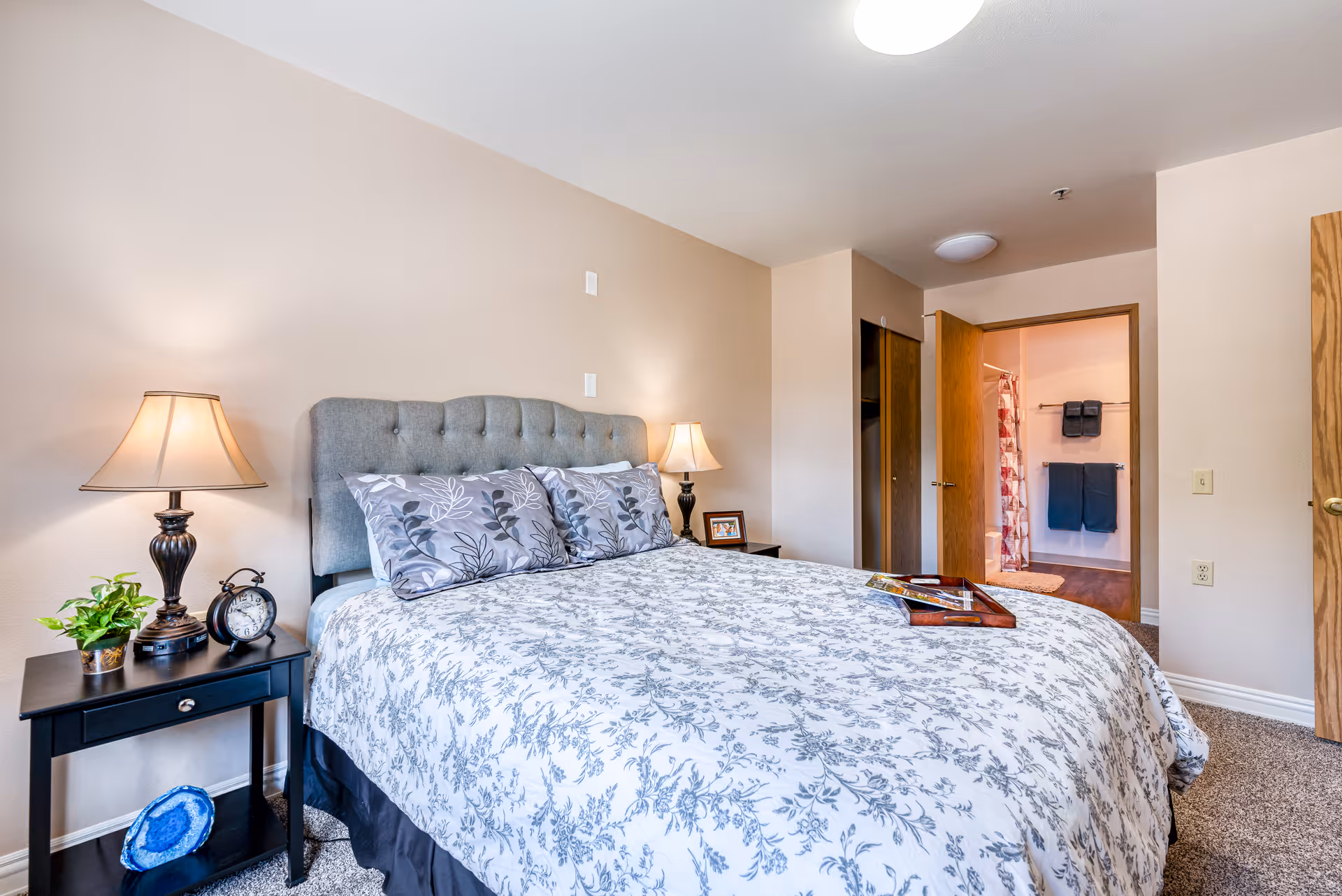 A cozy bedroom with a large bed featuring a gray tufted headboard and floral-patterned bedding. There are two matching pillows and a wooden tray on the bed. On the left side, there is a black nightstand with a table lamp, a small green plant, and an alarm clock. The room has beige walls and carpeted flooring. An open door reveals a bathroom with towels hanging on racks and a shower curtain with a red and white pattern.