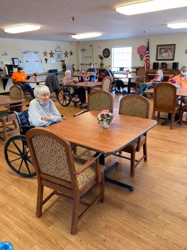 A group of elderly people sitting at tables in a nursing home dining or common area. Some are in wheelchairs, and others are seated in chairs. The room has wooden floors, decorated walls with stars and pictures, and an American flag in the corner. Each table has a small flower arrangement.
