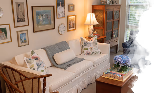 Cozy living room with a white sofa topped with floral pillows and a throw, framed artwork on the wall, a wooden cabinet and lamp, and a coffee table with flowers.