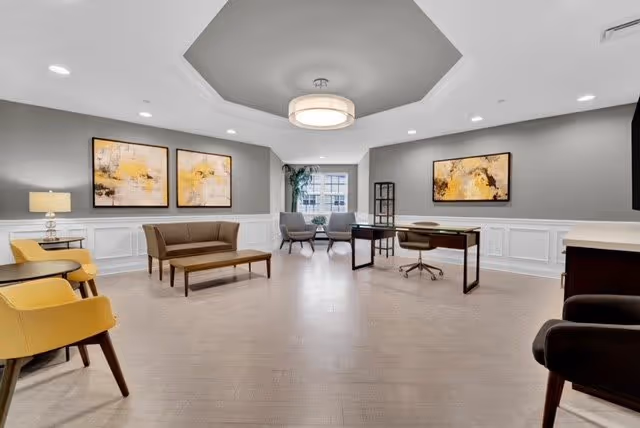 A spacious, well-lit common area with modern furniture including yellow and brown chairs, a wooden coffee table, a desk with a chair, and two gray armchairs near a window. The walls are painted gray with white wainscoting and decorated with abstract yellow and black artwork. The ceiling features a recessed design with a large circular light fixture.