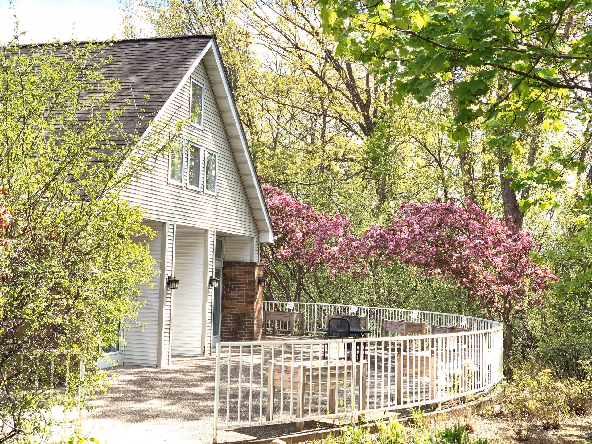 Outdoor patio area with white railing surrounding wooden benches and a grill, adjacent to a building with white siding and a steep roof. The area is surrounded by green trees and blooming pink flowers.