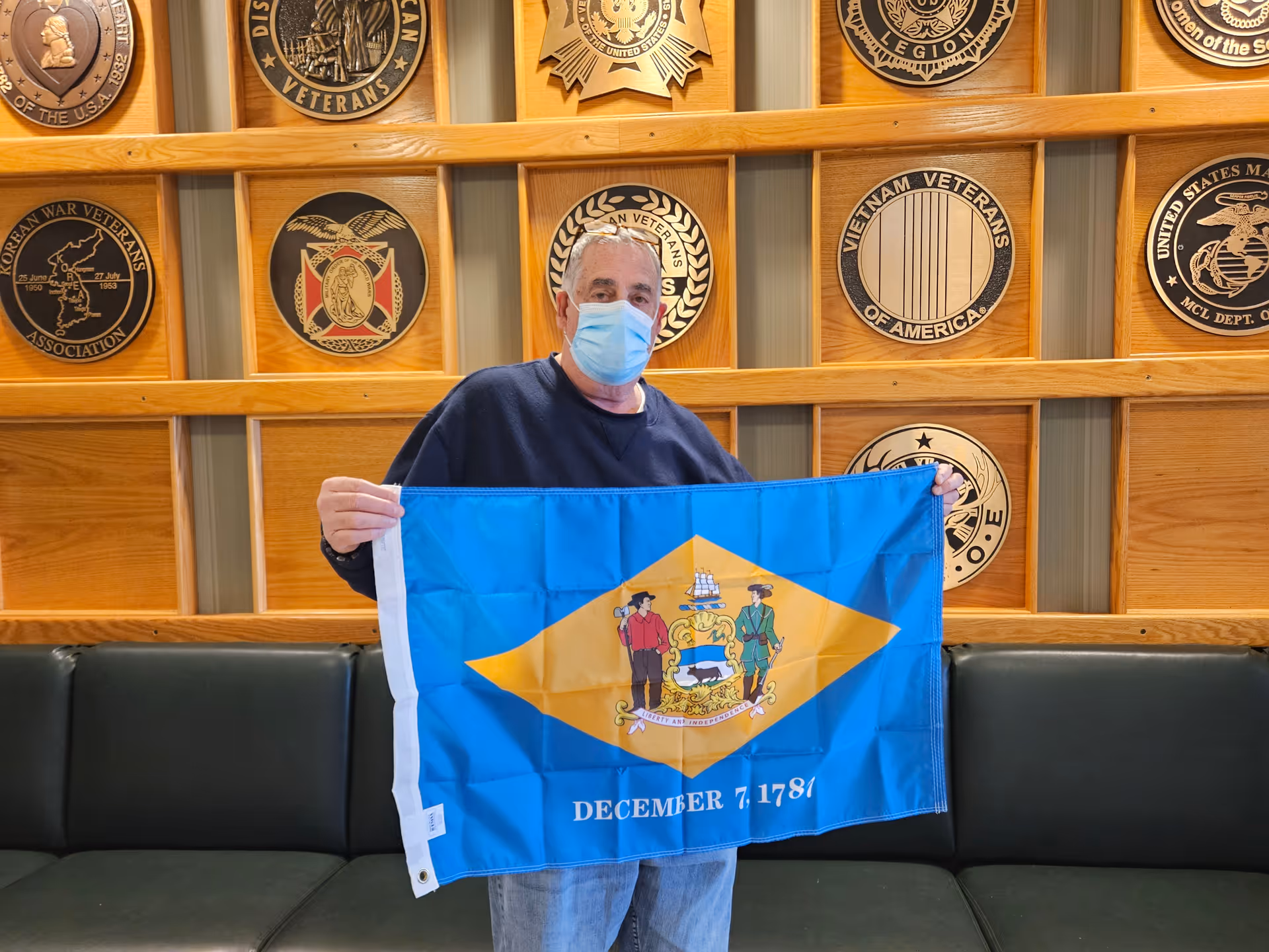 An elderly man wearing a face mask holds a Delaware state flag in front of a wooden wall decorated with various veterans' association plaques. He stands in a room with dark green cushioned seating along the wall.