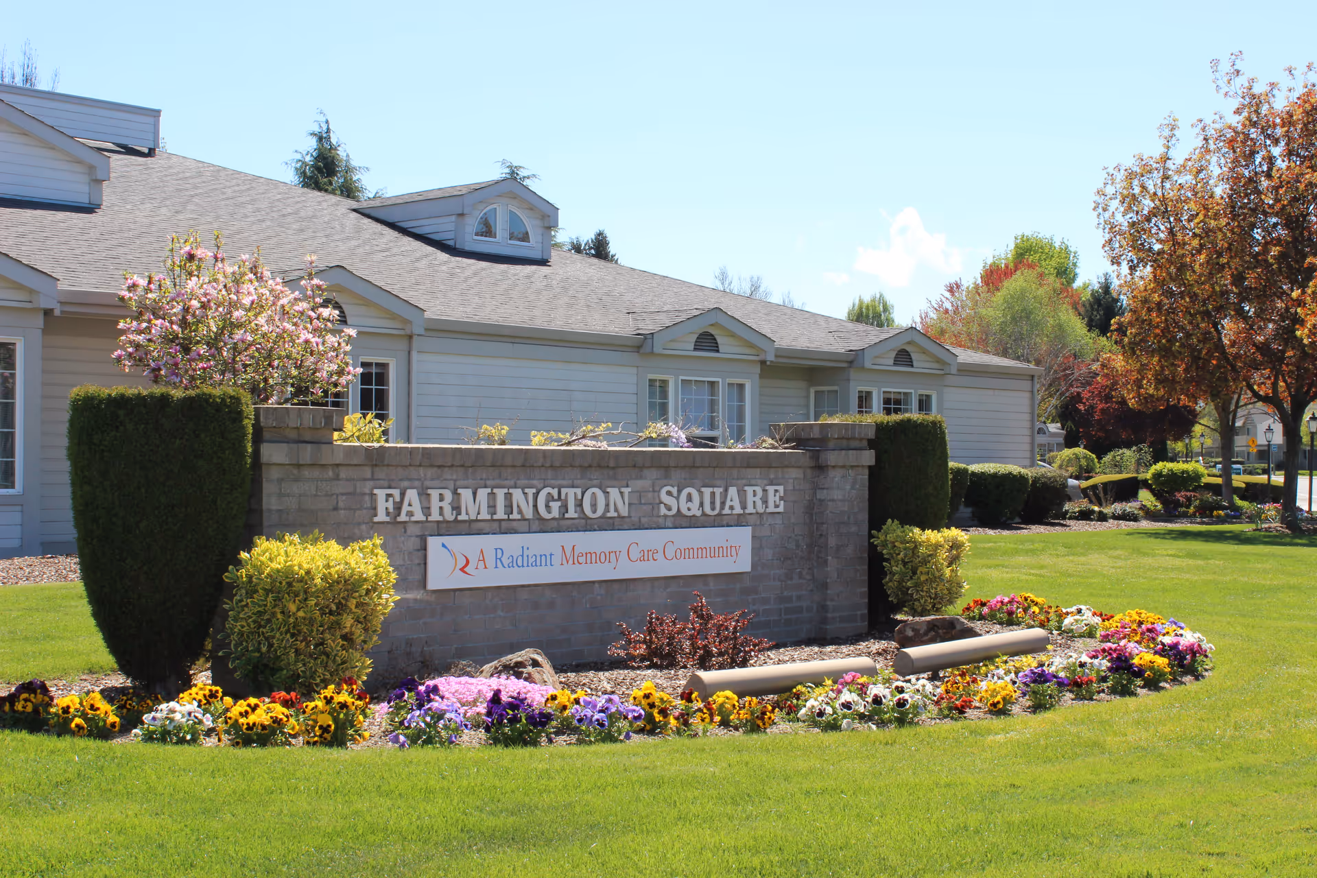 Exterior view of Farmington Square Medford facility with a brick sign displaying the name and tagline 'A Radiant Memory Care Community', surrounded by well-maintained green lawn, colorful flowers, trimmed bushes, and trees under a clear blue sky.