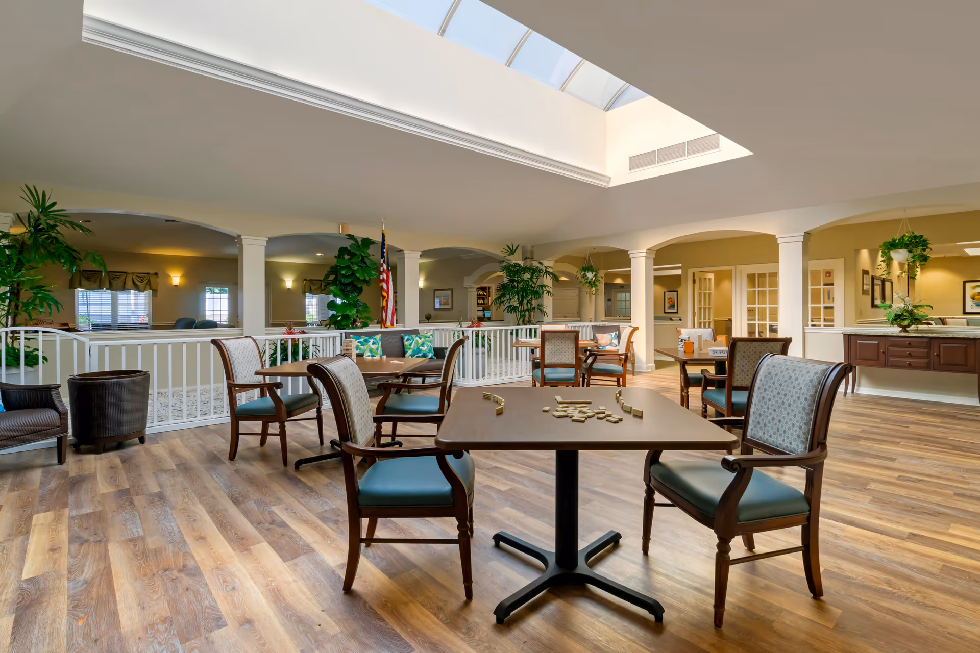 Spacious bright common lounge with tables and chairs, plants, and a skylight in a senior living facility.