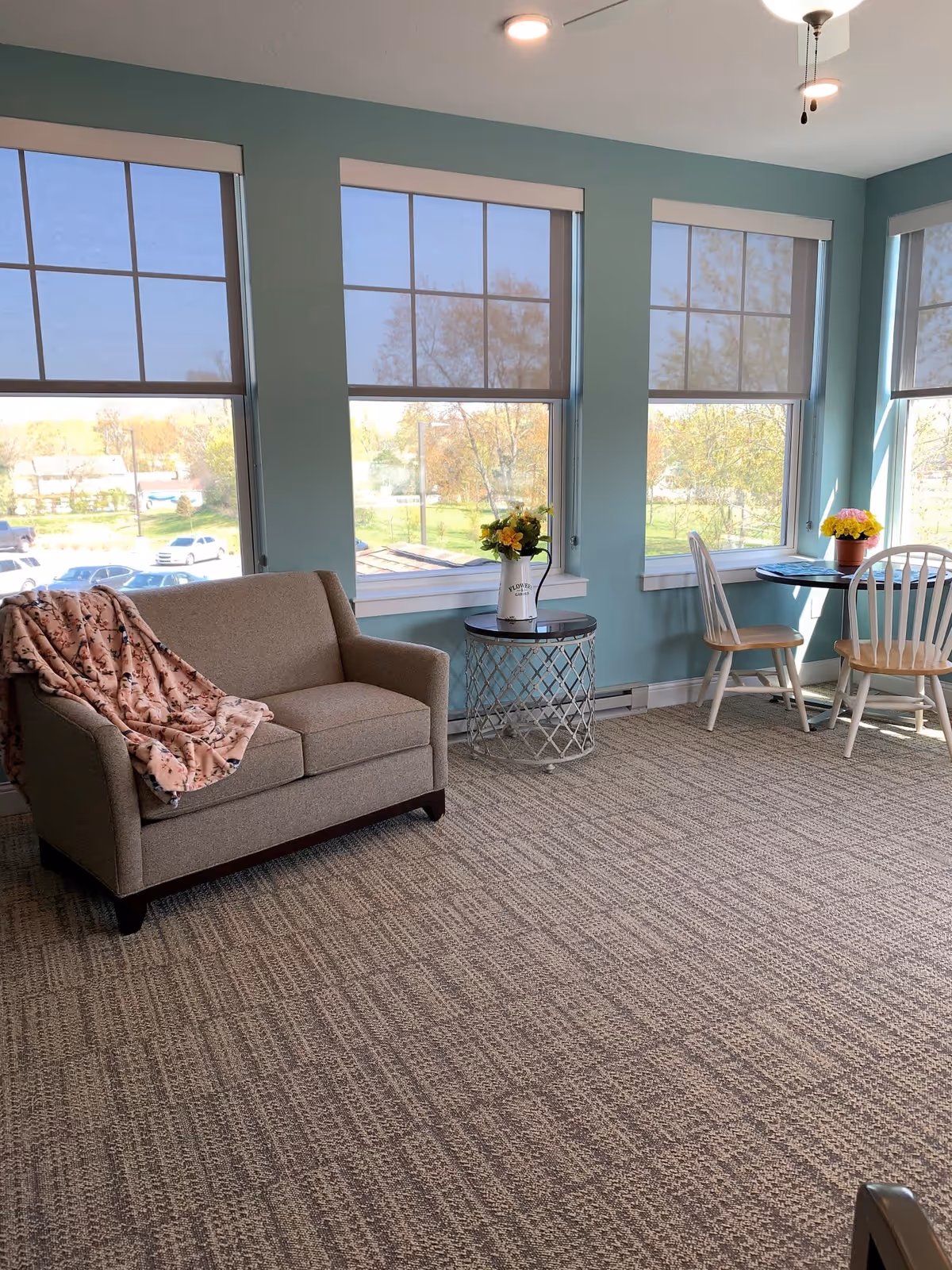 Bright sitting area with a loveseat, side table holding a vase of flowers, and a small round dining table with chairs in front of large windows.