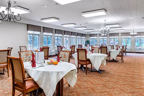 A spacious dining room with multiple round tables covered with white tablecloths, each set with folded red napkins, cups, and small decorative items. The room has large windows along one wall letting in natural light, chandeliers and ceiling lights, and patterned carpet flooring.