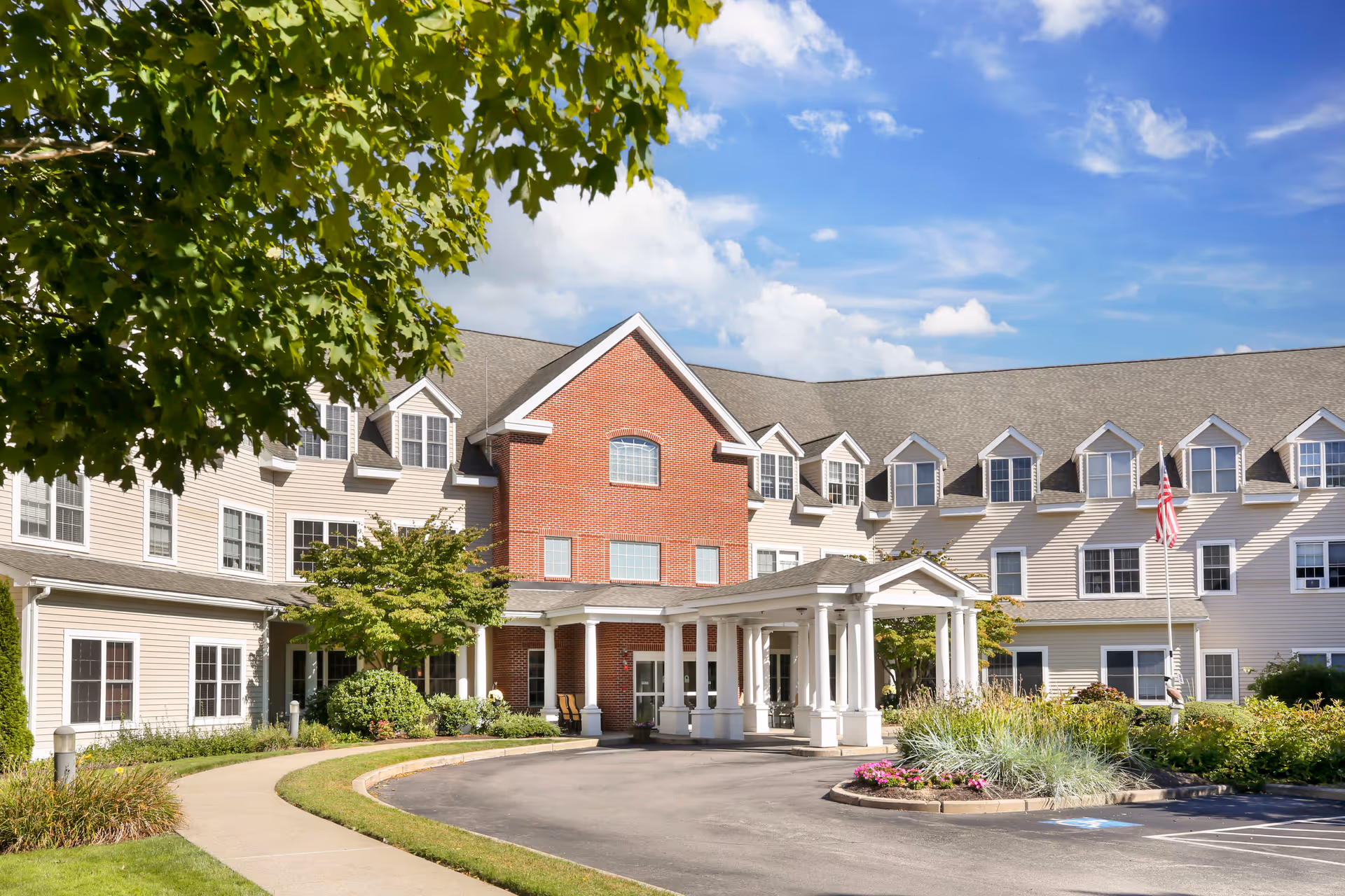 Exterior view of a senior living facility building with beige siding and a central red brick section. The building has multiple windows and dormers, a covered entrance with white columns, landscaped greenery, and an American flag on a flagpole. The sky is blue with some clouds.