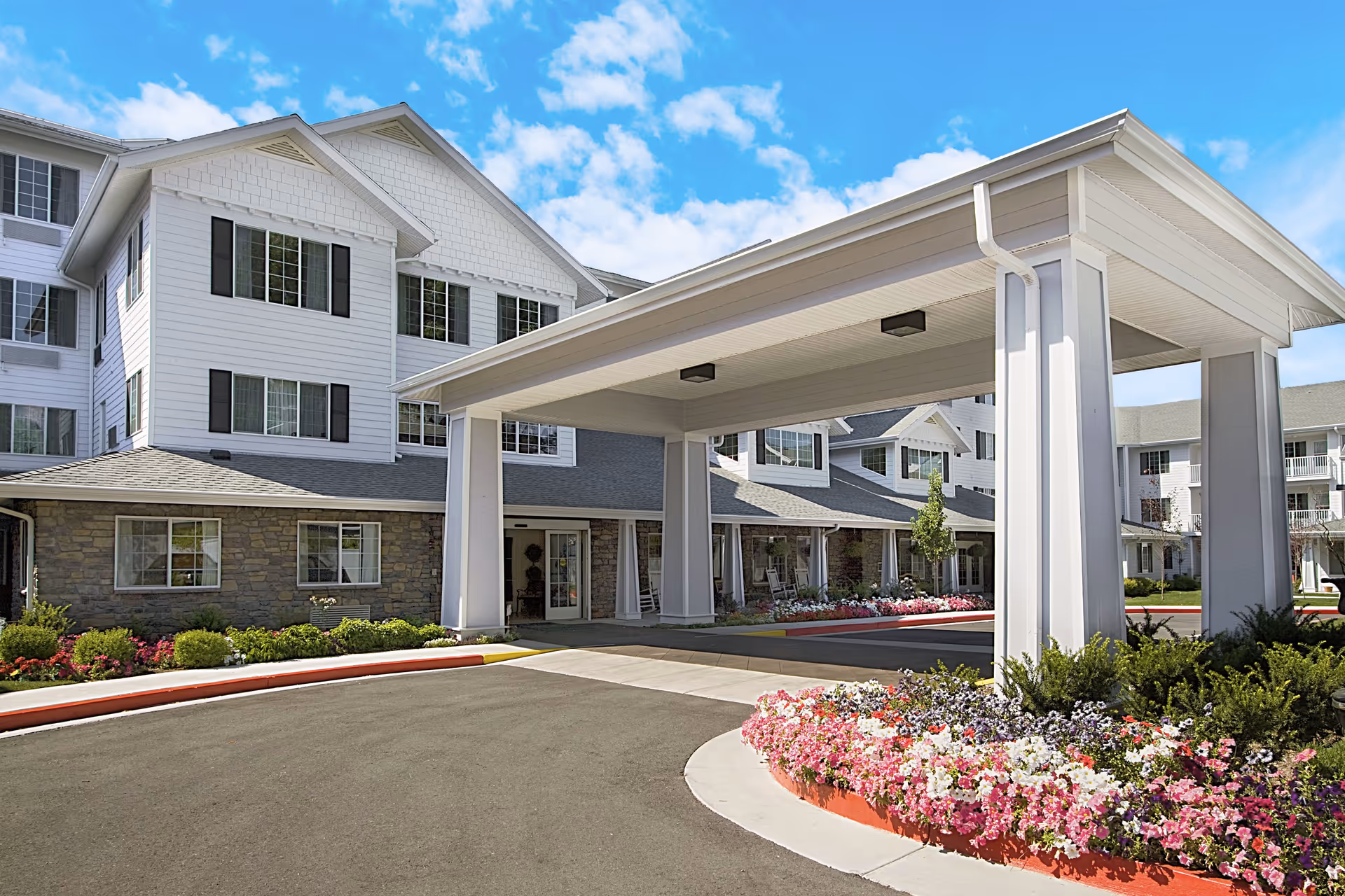 Exterior view of a multi-story senior living facility with white siding and stone accents. The building features large windows and a covered entrance with white pillars. There are colorful flower beds and neatly trimmed bushes along the driveway under a bright blue sky with scattered clouds.