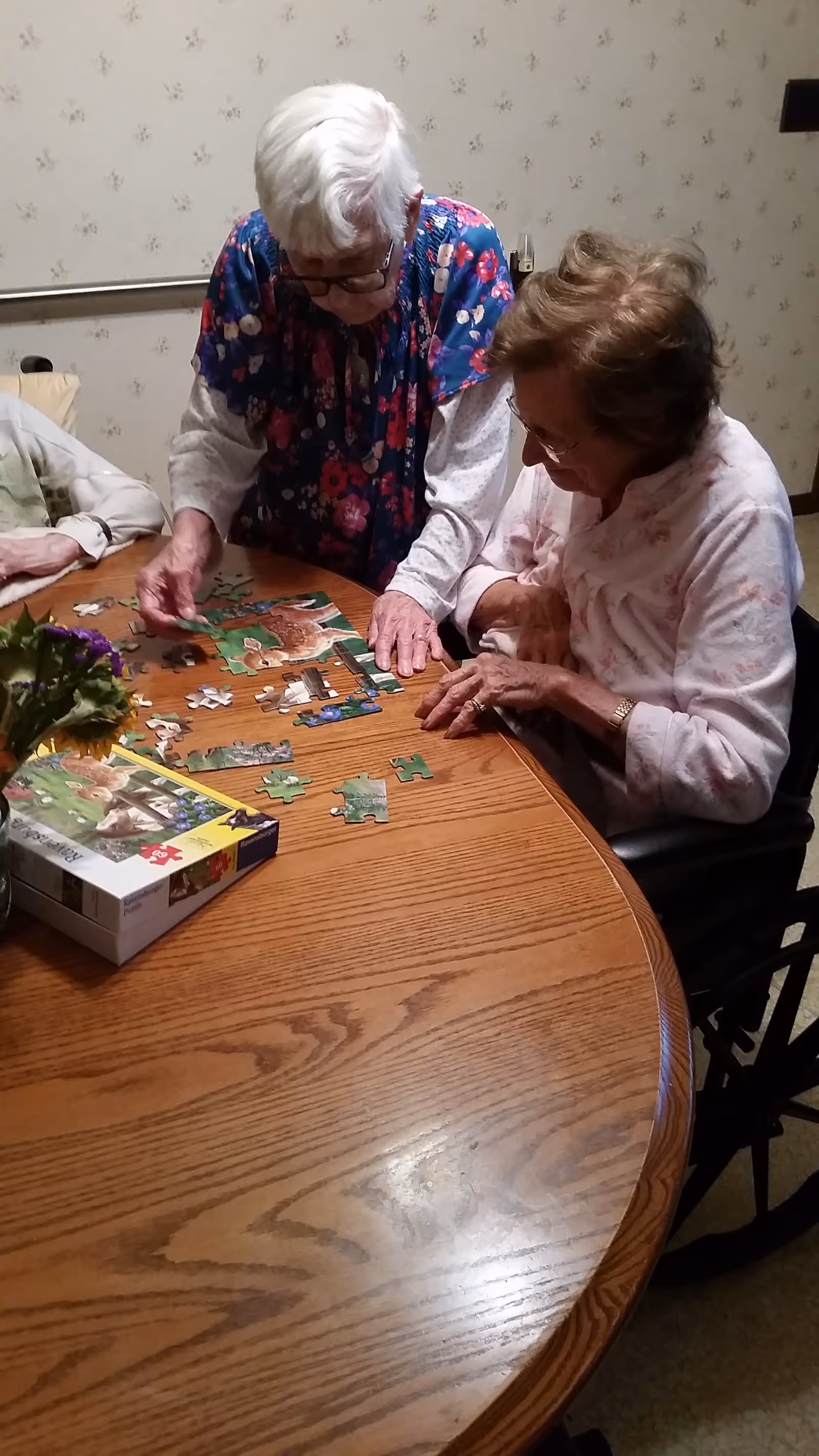 Two elderly women sitting at a wooden table working together on assembling a jigsaw puzzle in a room with light-colored wallpaper. One woman is standing and placing a puzzle piece, while the other is seated and watching attentively.