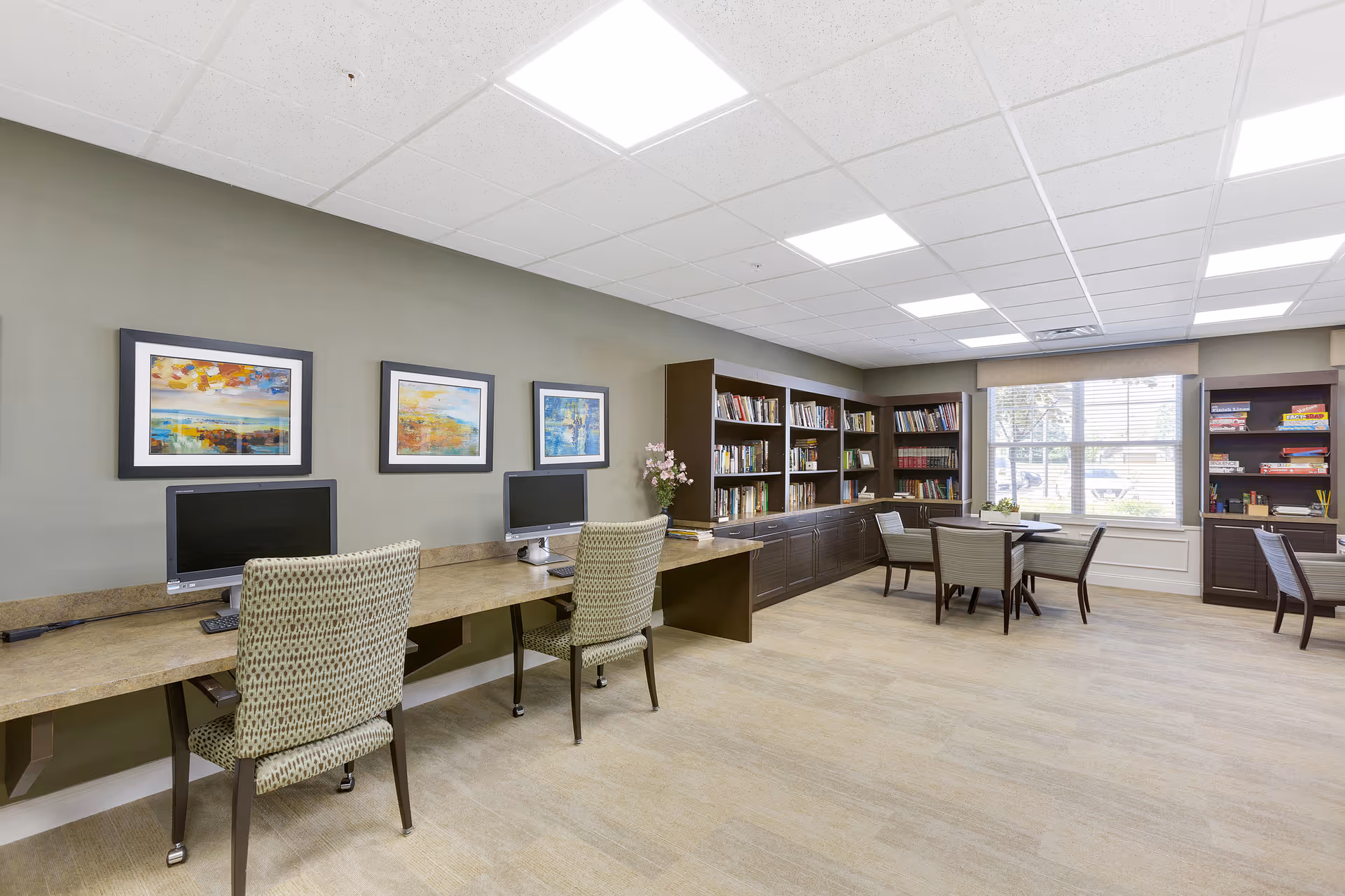 A well-lit room with a long desk featuring two computer stations with chairs. The walls are decorated with three framed abstract paintings. Along the back wall, there are bookshelves filled with books and board games. Near a large window with blinds, there is a round table surrounded by four chairs, creating a comfortable reading or activity area.