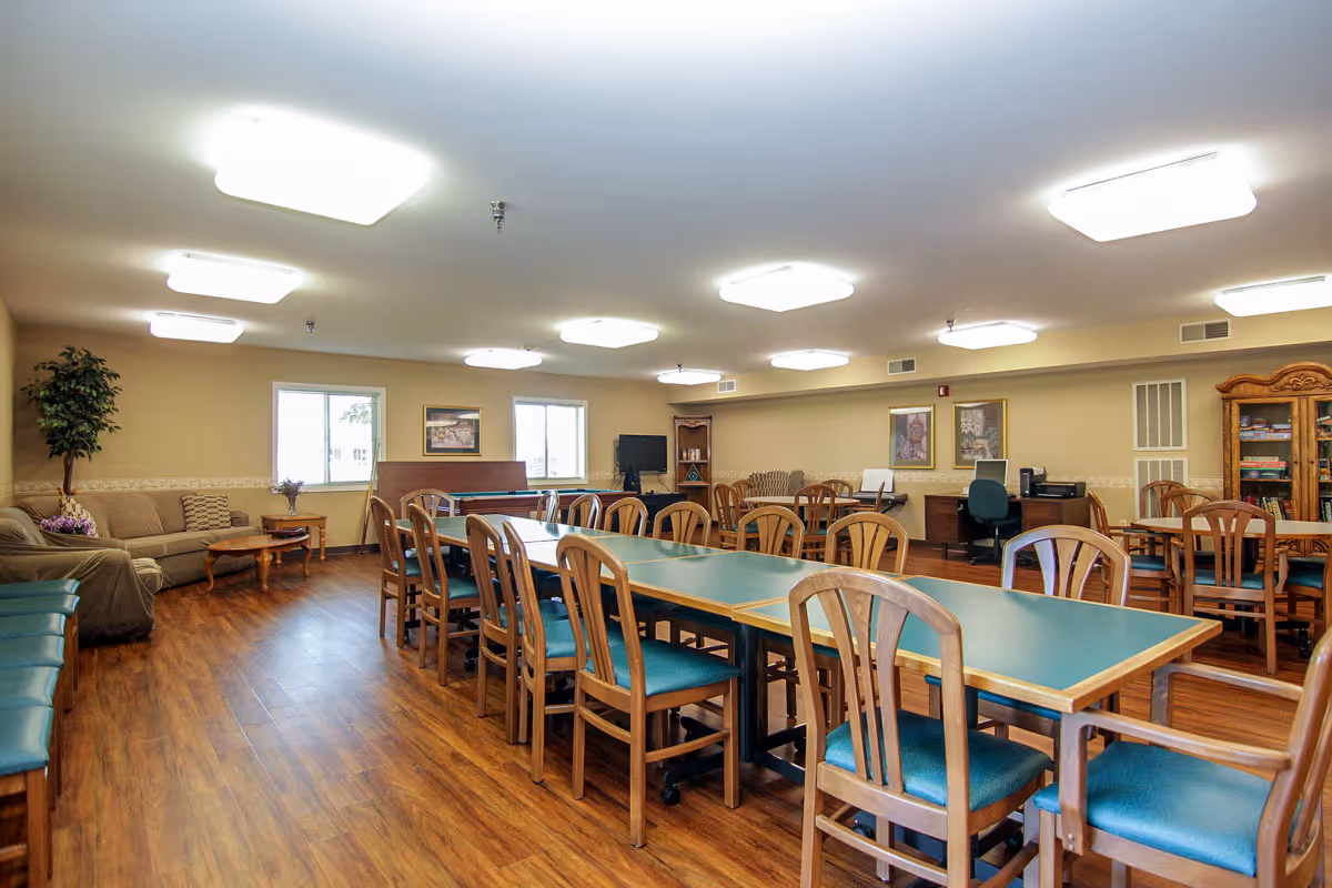 A spacious common room with multiple wooden tables and chairs arranged for group seating. The room features wood flooring, beige walls, several ceiling lights, a sofa with a coffee table, a TV, and bookshelves. There are windows letting in natural light and framed artwork on the walls.
