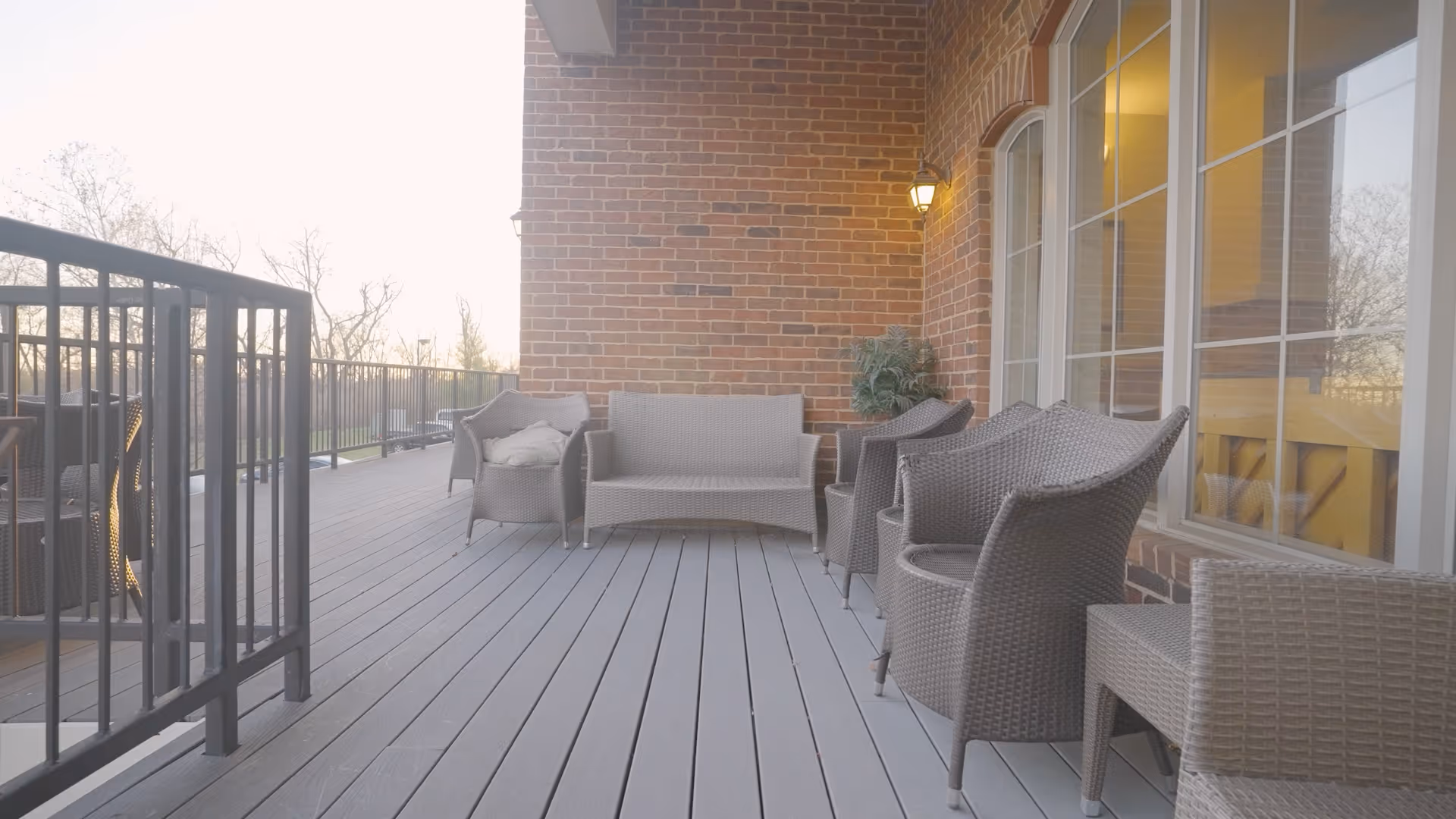 Outdoor patio area with gray wicker chairs and a loveseat arranged along a brick wall with large windows. There is a black metal railing on the left side and a small potted plant near the wall light.