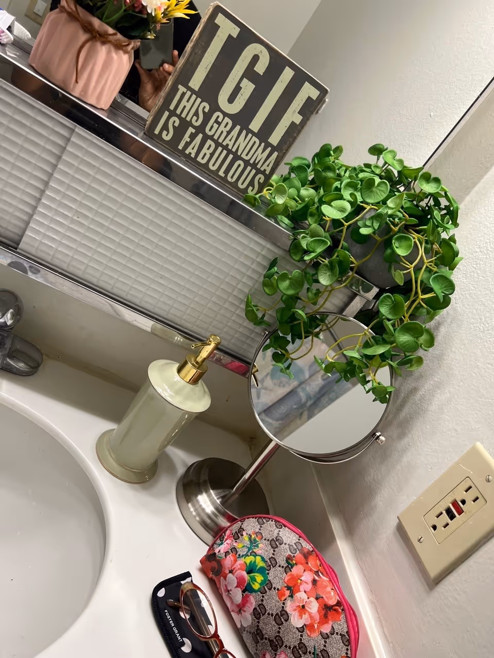 Bathroom sink countertop with a soap dispenser, small round vanity mirror, hanging faux plant, a sign reading 'TGIF THIS GRANDMA IS FABULOUS', and a floral makeup bag.
