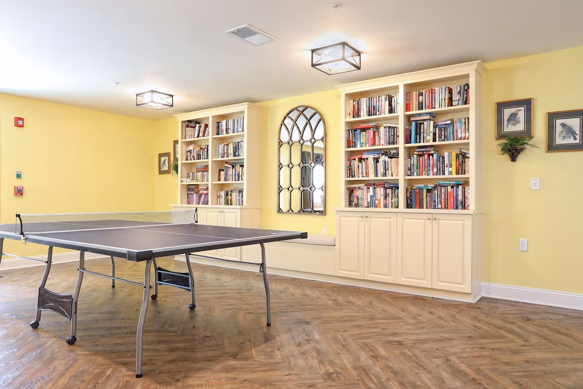 A bright common room with a ping-pong table on wood floors in front of built-in bookshelves and a decorative arched mirror.