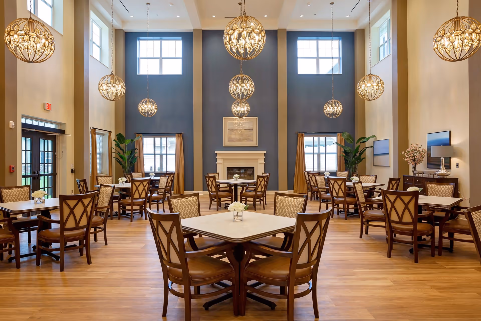 Spacious, well-lit dining room with multiple tables and chairs, hanging globe chandeliers, and a central fireplace.
