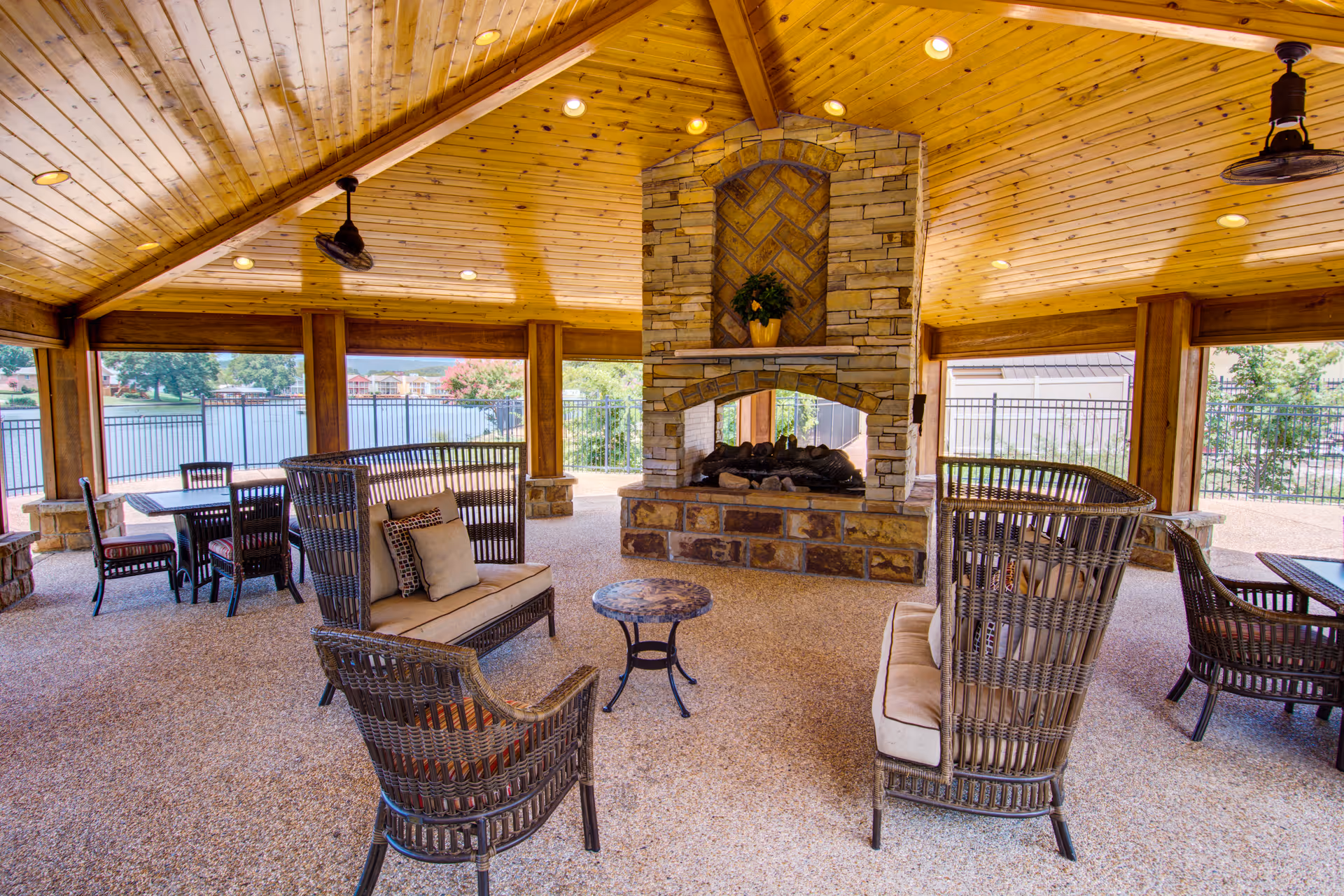 Covered outdoor lounge with wicker seating arranged around a central stone fireplace under a wood-paneled vaulted ceiling overlooking a lake.