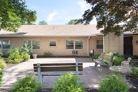 Outdoor courtyard area of a nursing and rehabilitation center with two wooden benches facing each other, surrounded by greenery and trees, with a beige brick building in the background under a partly cloudy sky.