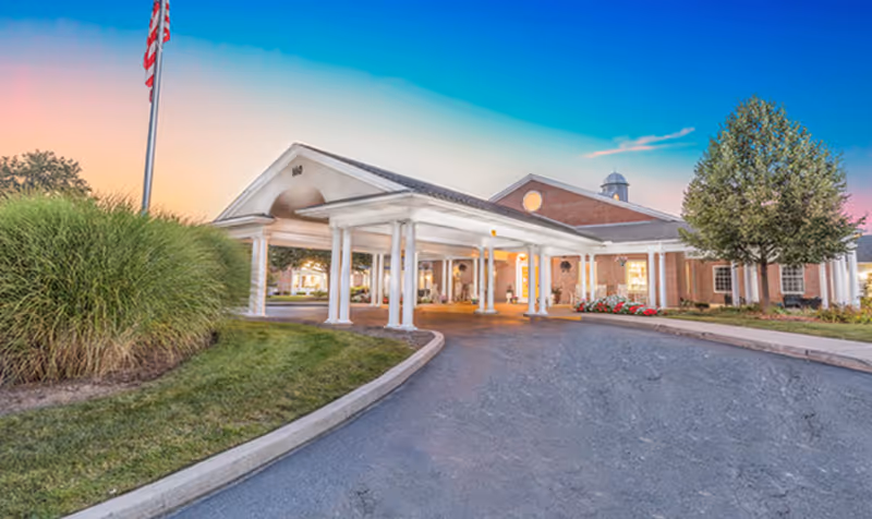 Exterior view of a senior living facility building with a covered entrance supported by white columns, a flagpole with an American flag, well-maintained landscaping including grass, bushes, and a tree, under a clear sky at sunset.