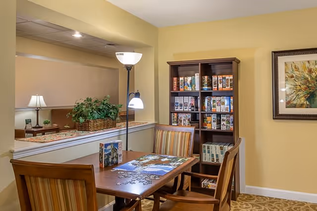 A cozy interior room with a wooden table and four striped cushioned chairs. On the table, there is a partially completed jigsaw puzzle and puzzle boxes. Behind the table, there is a tall wooden bookshelf filled with various board games and puzzles. The walls are painted light yellow, and there is a framed floral artwork hanging on the right wall. A floor lamp with two lights stands next to the table, and a half wall with a plant basket separates this area from another room with a table lamp and additional seating.