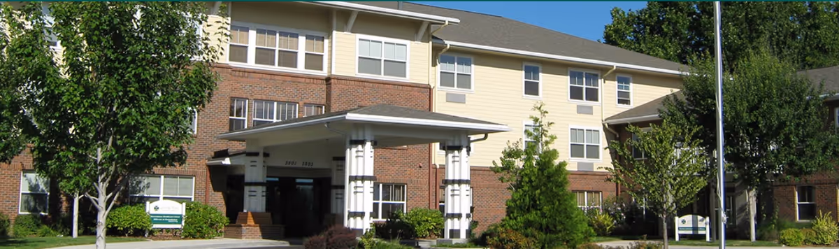 Front exterior of a three-story senior living building with a covered entrance, brick and siding facade, and landscaped trees.