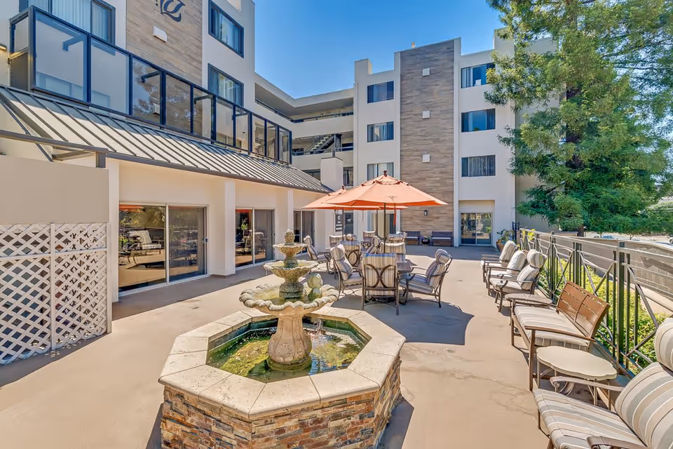 Outdoor patio area at Burlingame Senior Living featuring a stone fountain in the foreground, surrounded by cushioned chairs and tables with an orange umbrella. The patio is adjacent to a multi-story building with large windows and a tree providing greenery on the right side.