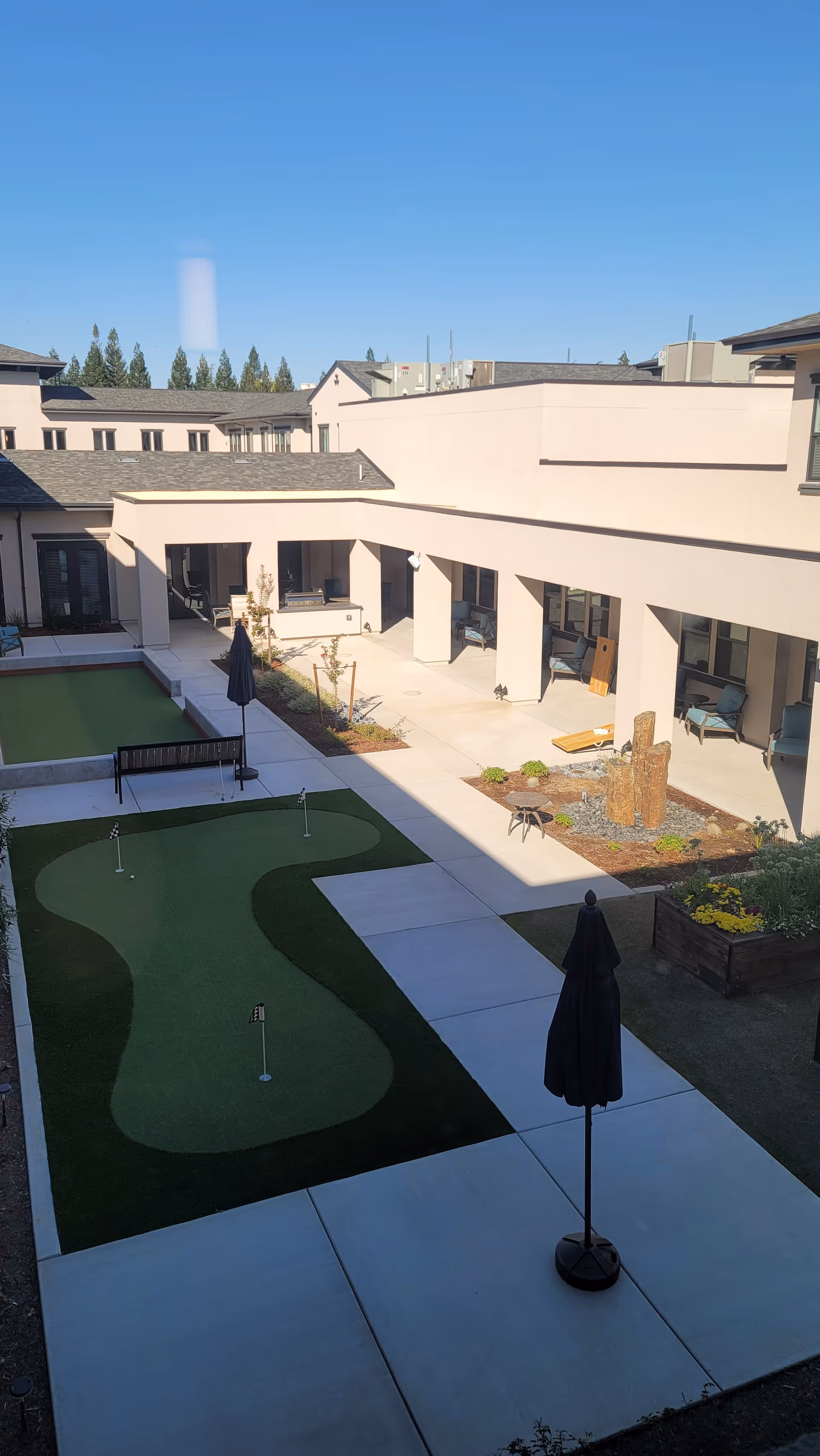 Courtyard of a senior living facility with small putting greens, paved walkways, umbrellas, seating areas and surrounding two-story building under a clear blue sky.