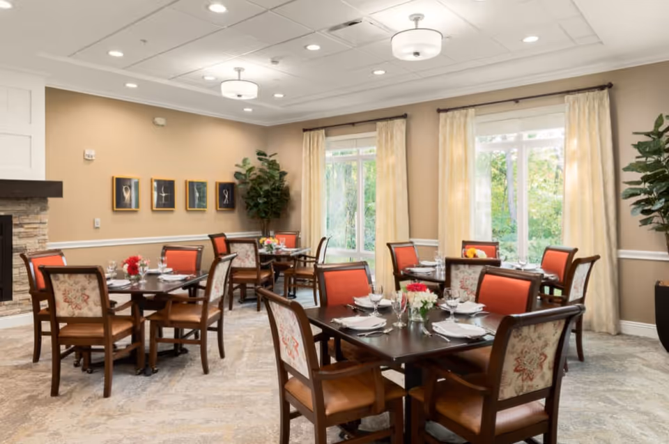 A bright dining room with several dark wood tables set with white plates, glasses, and silverware. Each table has a small floral centerpiece. The chairs have wooden frames with floral upholstery on the backrests and orange cushions. Large windows with cream-colored curtains allow natural light to fill the room. The walls are beige with white trim, and there is a stone fireplace on the left side. Four framed artworks hang on the wall above the fireplace, and a large potted plant is in the corner near the windows.