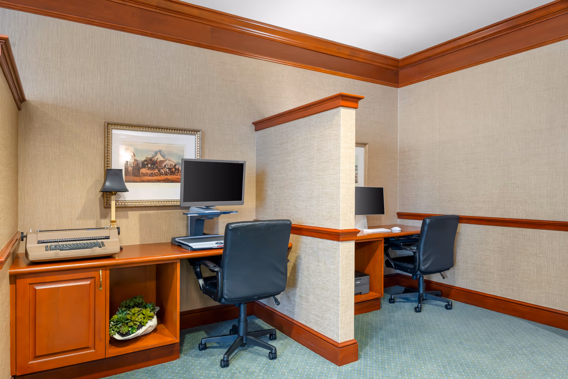 Two wood-trimmed computer workstations with monitors, black swivel chairs, and a decorative print in a carpeted business center.