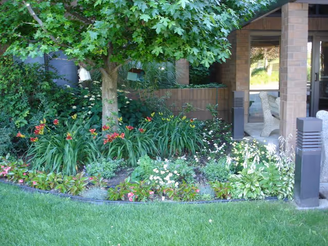 A landscaped garden area with a variety of green plants and colorful flowers surrounding a tree. In the background, there is a covered patio area with stone benches and brick pillars.