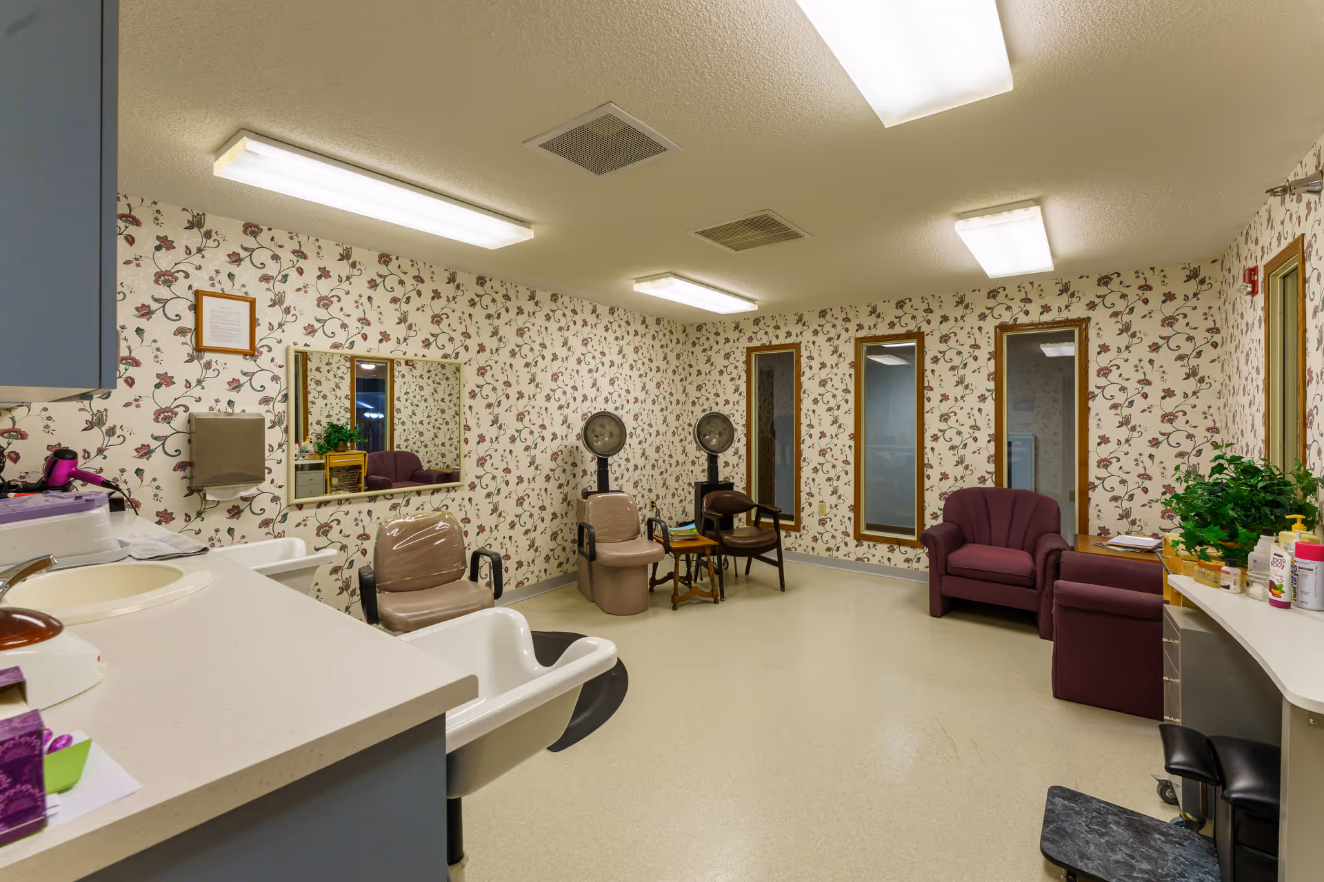 Interior of a hair salon or beauty room with floral wallpaper, two hair drying chairs with hooded dryers, a purple armchair, a counter with a sink, and various hair care items and plants on the counters.