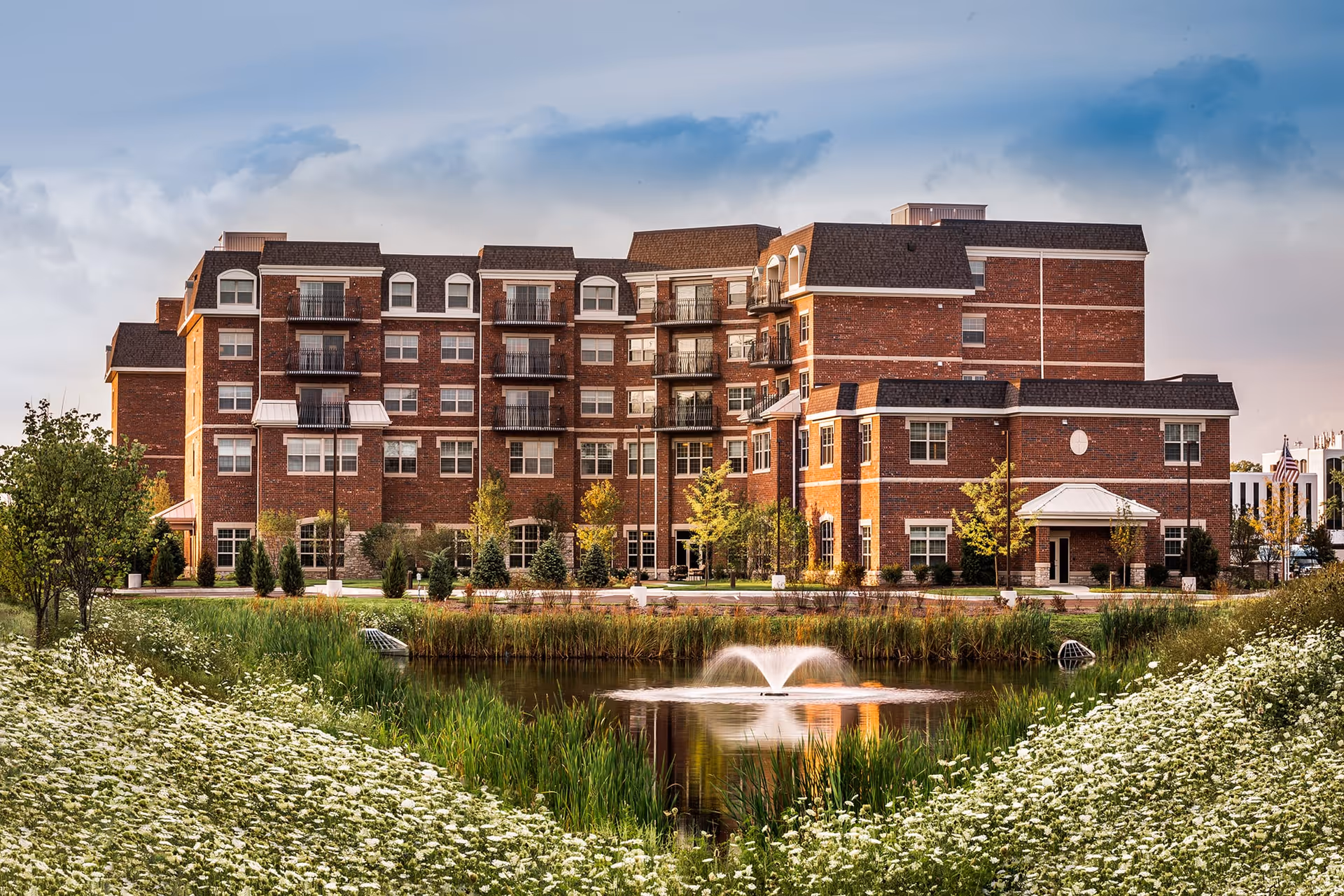 A large multi-story brick building with multiple balconies, surrounded by landscaped greenery and trees. In the foreground, there is a pond with a water fountain, bordered by white flowers and tall grass under a partly cloudy sky.