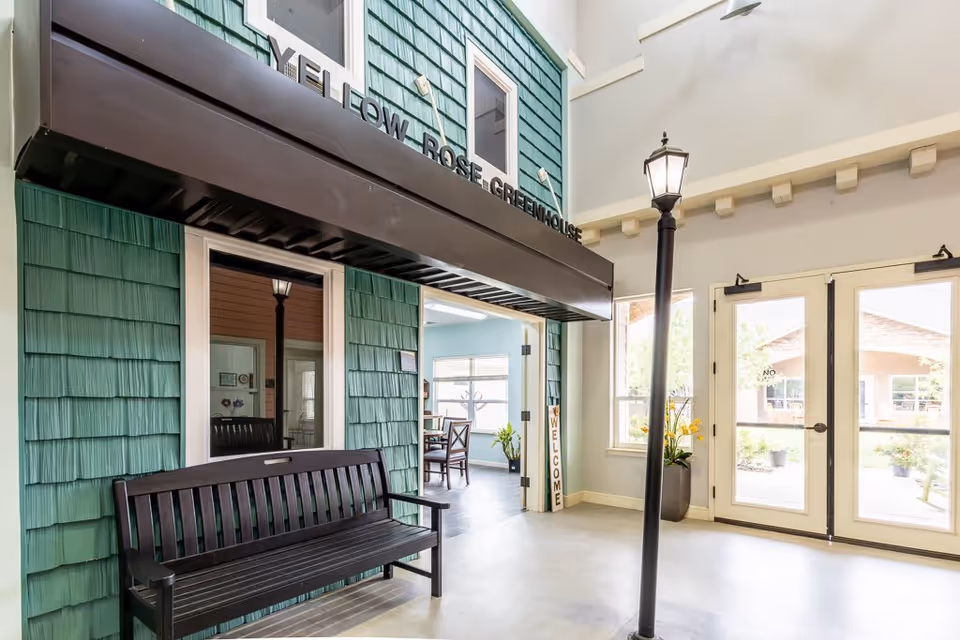 Interior view of a senior living facility area with green shingle siding, a black bench, a tall black lamp post, and double glass doors leading outside. A sign above the bench reads 'YELLOW ROSE GREENHOUSE'. There is a 'WELCOME' sign leaning against the wall near the open doorway leading to a room with tables and chairs.