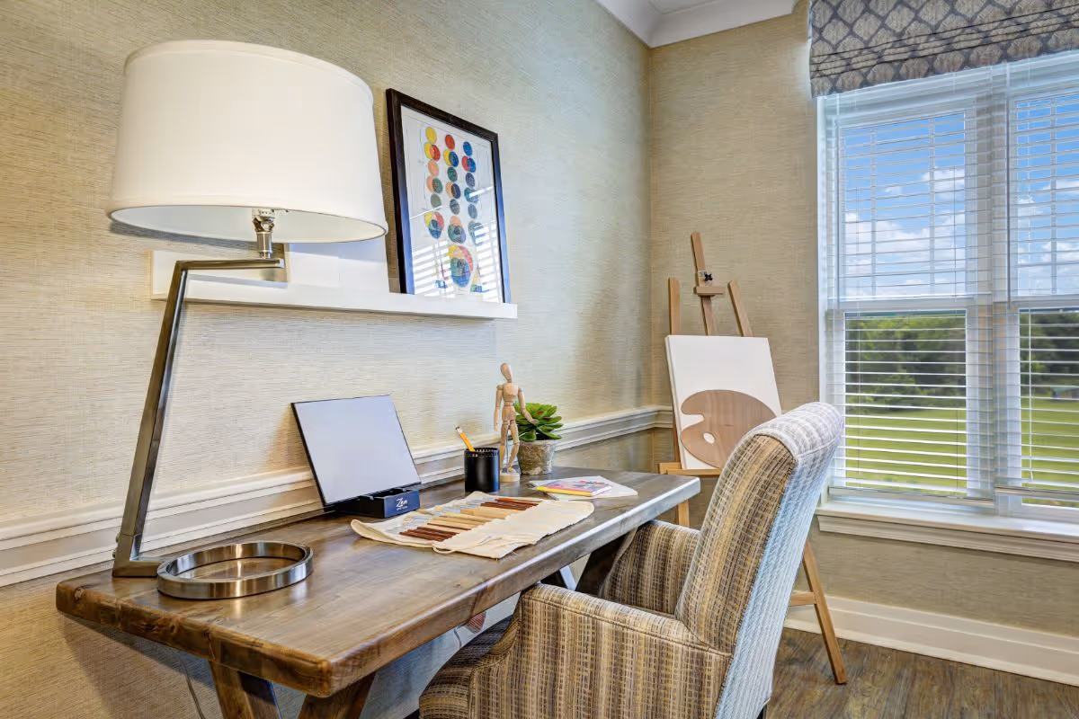 A bright study corner with a wooden desk, modern lamp, upholstered chair, art supplies and a window with blinds overlooking a lawn.