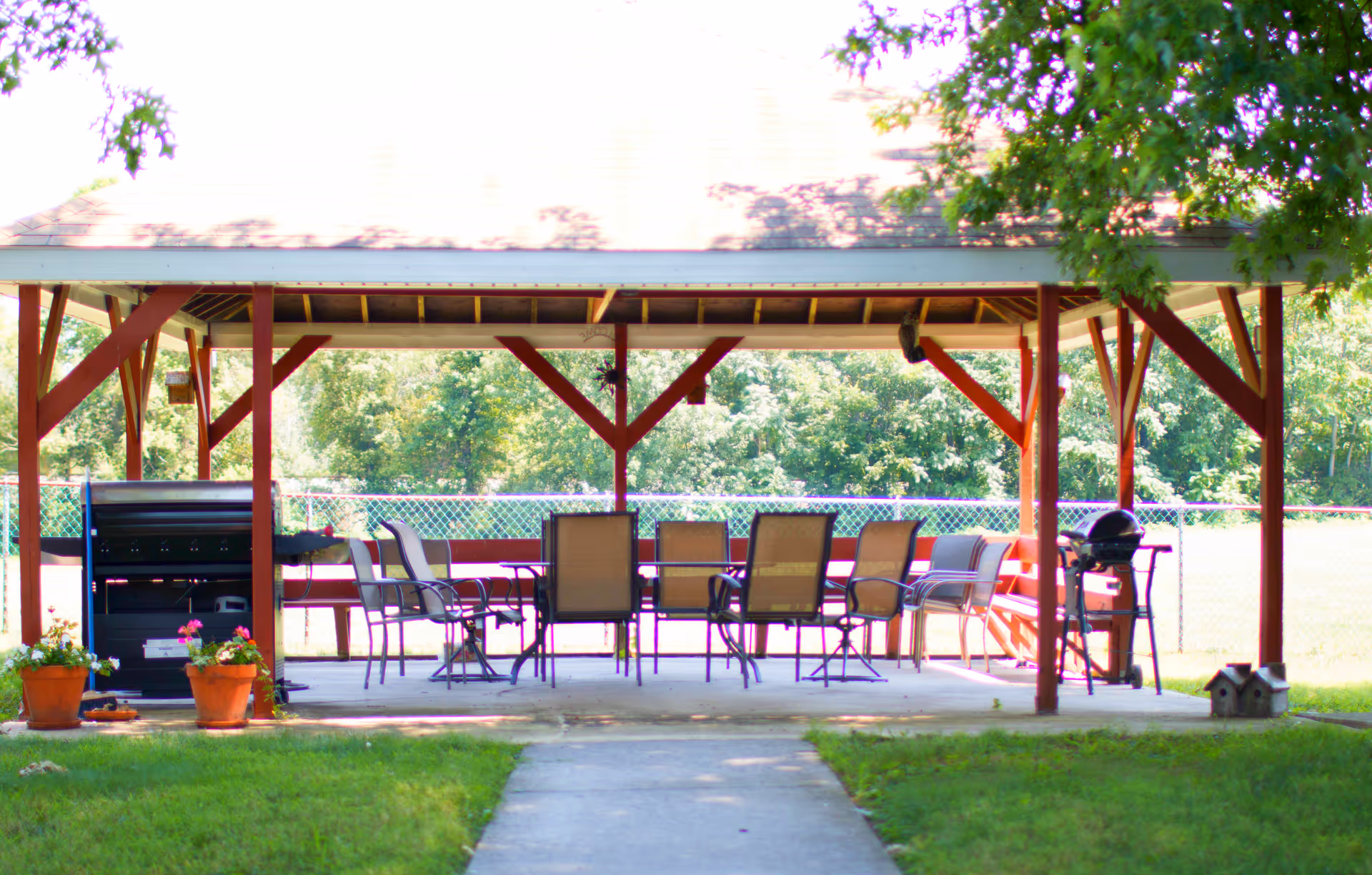 Outdoor covered pavilion with a roof supported by red wooden beams, containing several chairs and tables arranged for seating. There are two grills on either side of the pavilion and potted plants placed near the entrance. The area is surrounded by green grass and trees in the background.