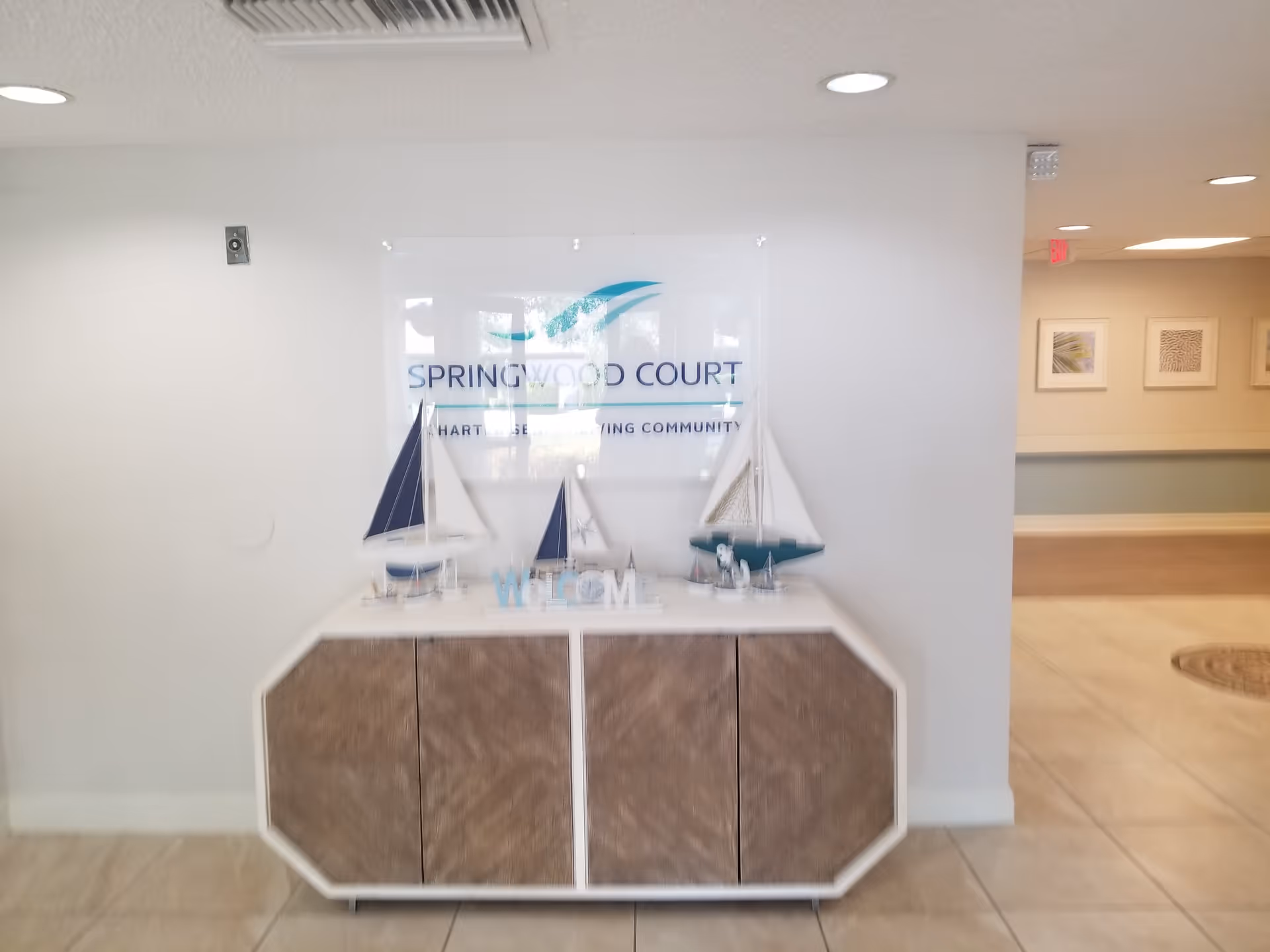 Interior view of a senior living facility lobby area with a white and wood cabinet displaying decorative model sailboats and a welcome sign. Behind the cabinet is a wall-mounted sign that reads 'Springwood Court'. The floor is tiled, and the walls are painted white with some framed artwork visible in the hallway to the right.