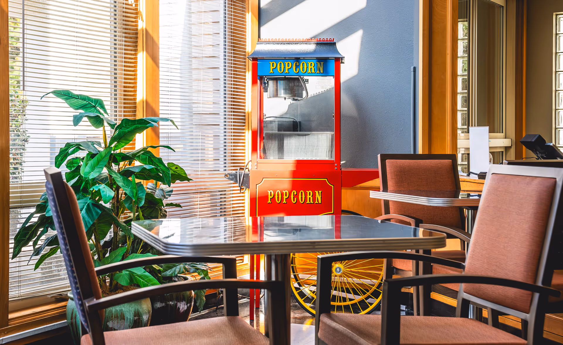 A bright indoor seating area with a metal table and four cushioned chairs. Behind the table is a vintage-style red popcorn machine with yellow wheels and the word 'POPCORN' written on it. To the left, there is a large green potted plant near a window with blinds, allowing sunlight to stream in.