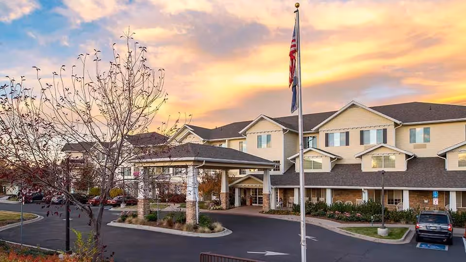 Front entrance of a senior living building with a covered porte-cochère, an American flag on a pole, and a landscaped driveway under a colorful sunset sky.