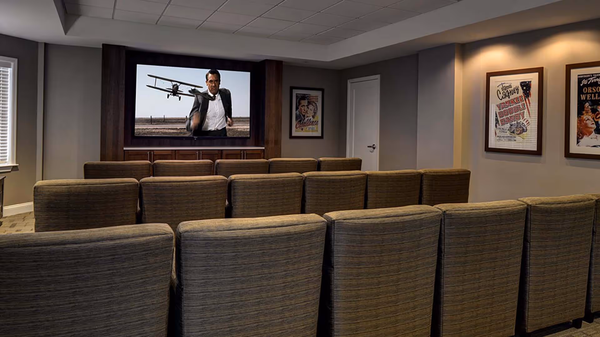 A small movie theater room with three rows of cushioned chairs facing a large screen showing a man running with a biplane in the background. The room has gray walls with framed vintage movie posters and a window with blinds on the left side.