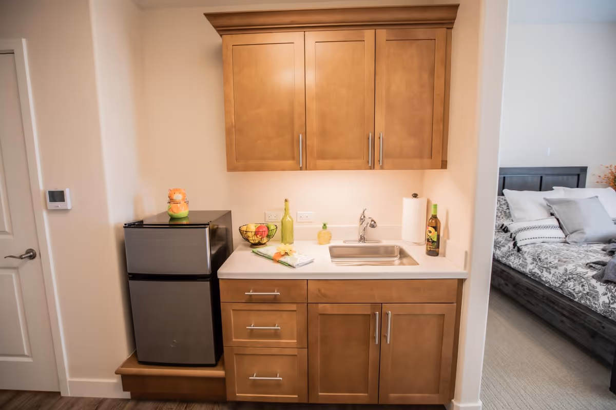 A small kitchenette area with wooden cabinets, a stainless steel mini refrigerator, a sink, and a countertop with a fruit basket, a bottle, and a paper towel holder. To the right, a bedroom with a bed, pillows, and a blanket is partially visible.