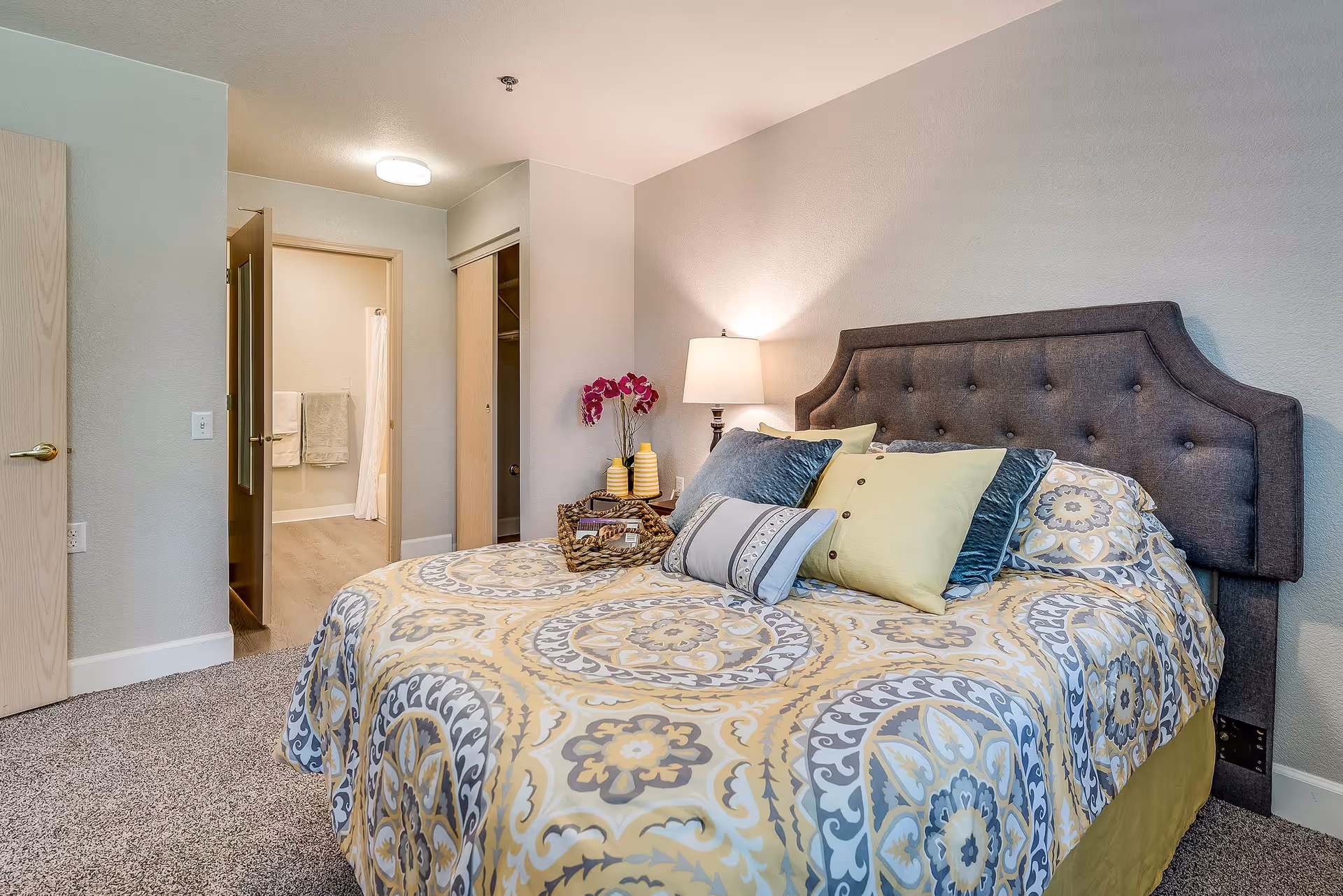 Well-decorated bedroom with a patterned bedspread, tufted headboard, bedside lamp and a view into an adjacent bathroom.