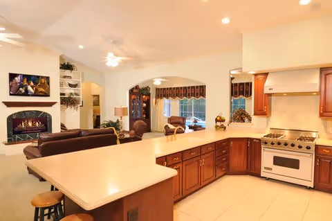 Interior view of a living space in Timberlane Lodge Assisted Living Facility featuring a kitchen with wooden cabinets, a white stove, and a large countertop island with stools. Adjacent to the kitchen is a living room area with brown leather sofas, a fireplace, and a wall-mounted TV. The space is well-lit with ceiling lights and fans.