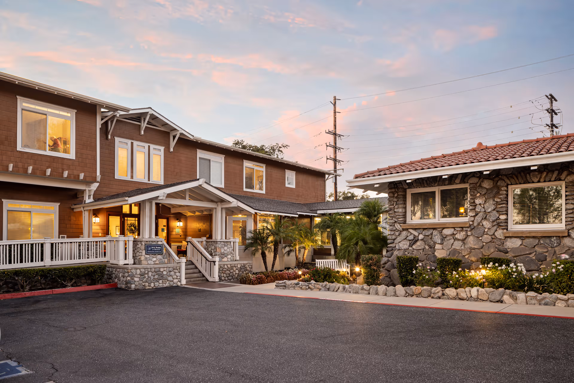 Exterior view of a senior living facility named Ivy Park at Claremont during sunset. The building features a combination of stone and wood siding with a tiled roof on one section. There is a ramp and stairs leading to the entrance, surrounded by well-maintained landscaping including bushes and palm trees. The sky is partly cloudy with a soft pink and blue hue.